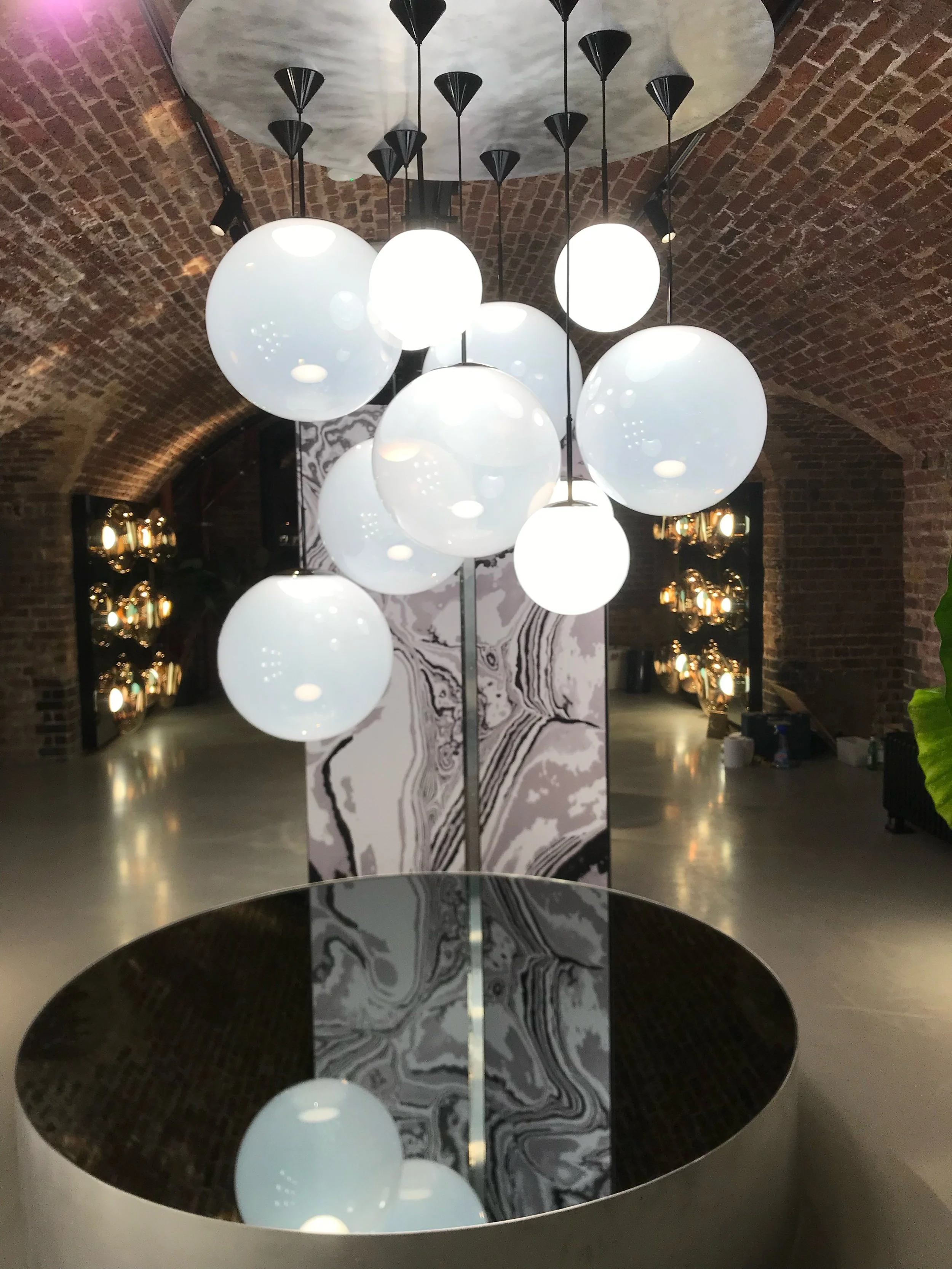 Modern chandelier with multiple white spherical lights hanging in an arched brick interior, with a large round mirror table below reflecting the lights.