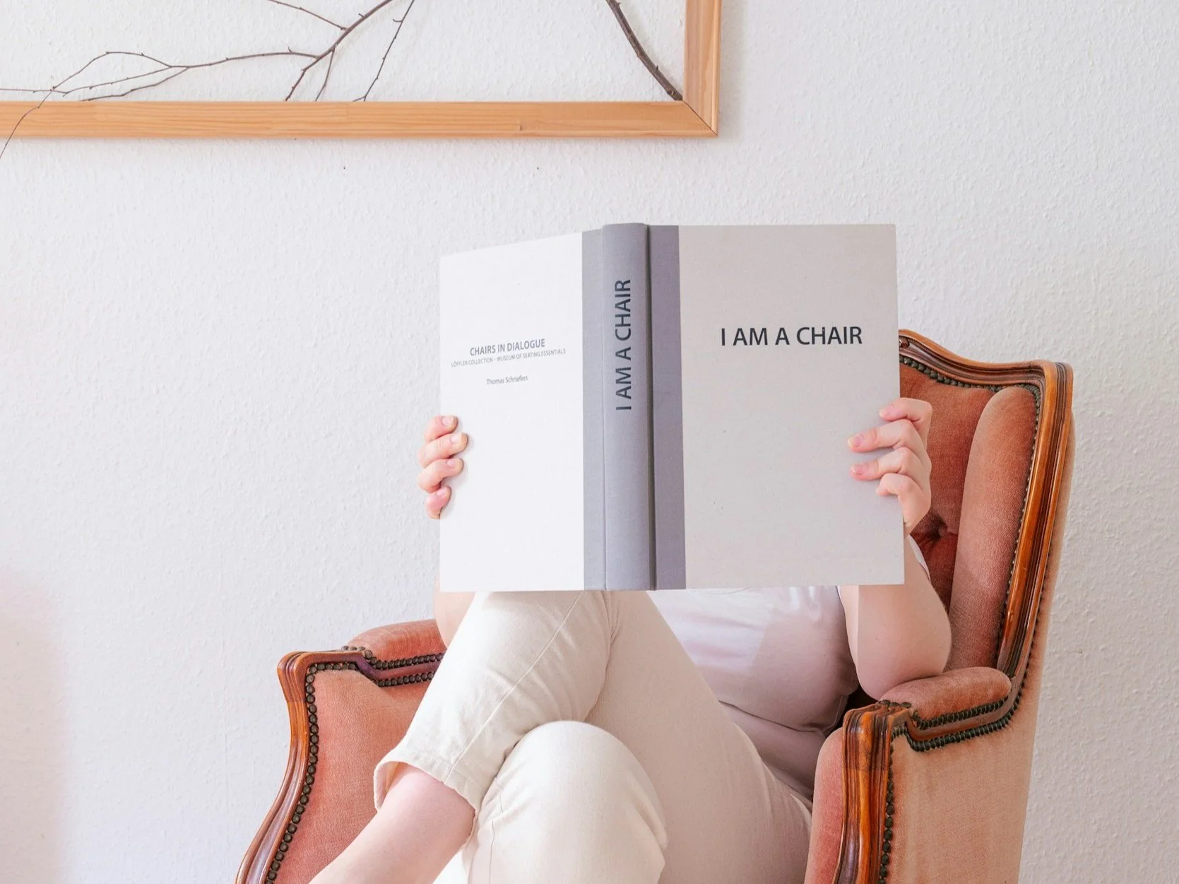 Person sitting in a vintage armchair, holding a large book titled 'I AM A CHAIR' in front of their face, with a white textured wall and framed artwork in the background.