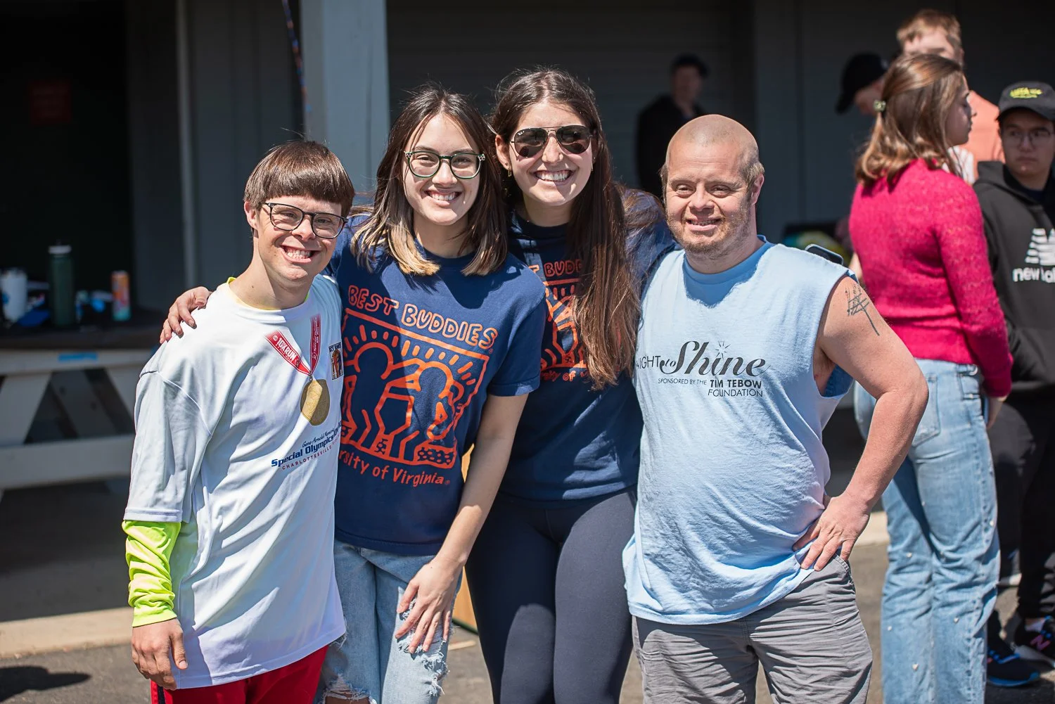 Four people smiling outdoors, posing for a photo. They are wearing casual clothing, some with event shirts. The group includes two women and two men, with one of the men having a tattoo on his arm. Other people are visible in the background.