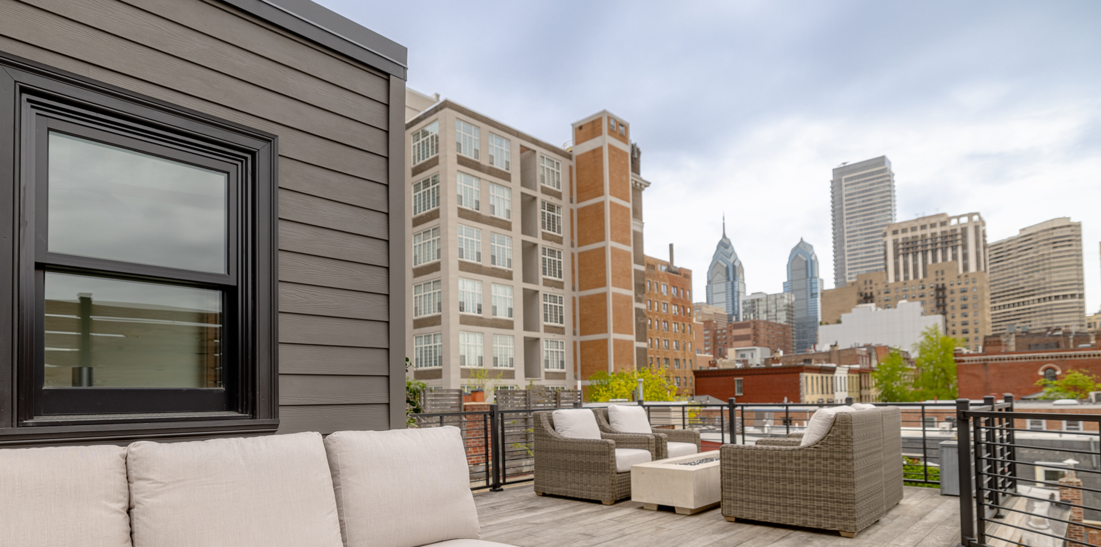 Outdoor roof top deck with beige cushioned seating and wicker chairs, surrounded by a black metal railing, with a city skyline of tall buildings and a cloudy sky in the background.