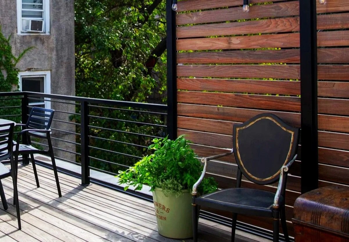 An outdoor balcony with a black railing, wooden floorboards, a wooden privacy screen, two black plastic chairs, a potted plant, and a black and gold decorative chair. There are green trees and an apartment building with windows in the background.