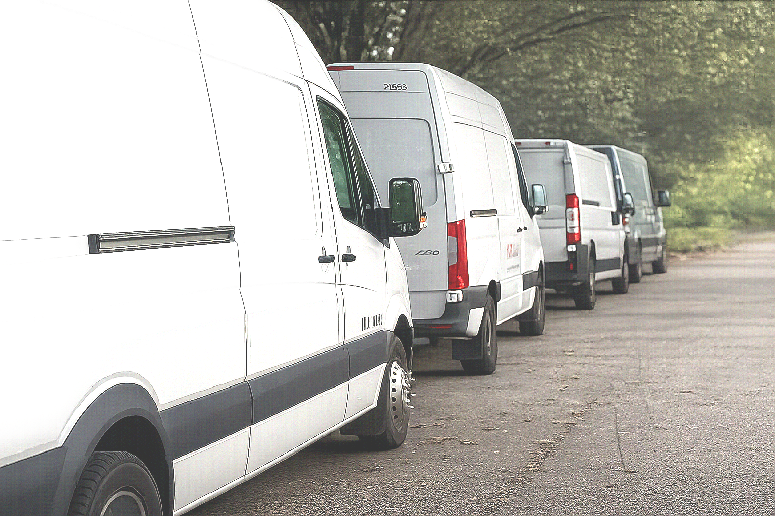A line of white cargo vans parked on the side of a paved road with trees in the background.