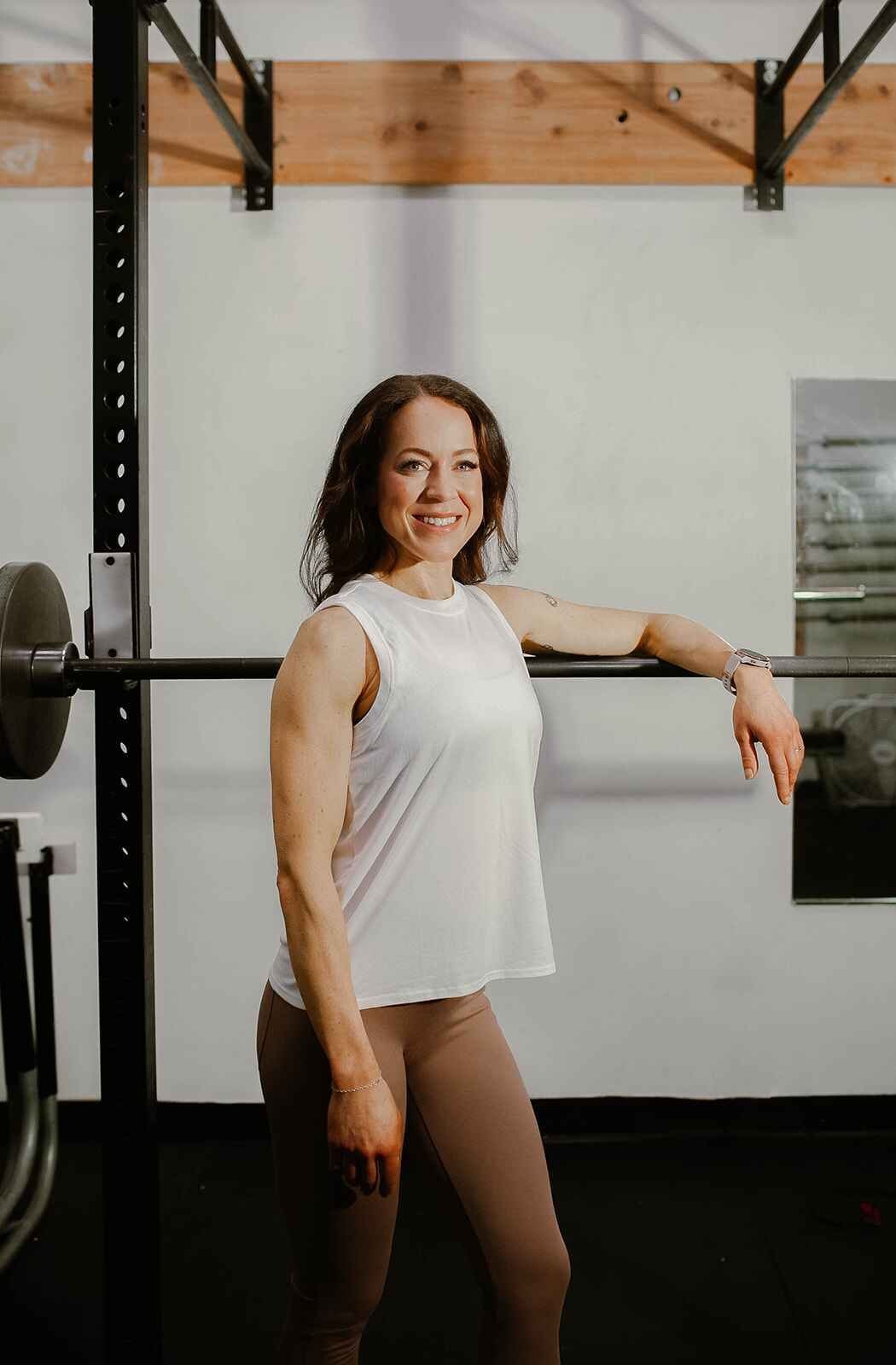 A woman standing in a gym near a barbell with a smile, wearing a white sleeveless top and beige leggings.