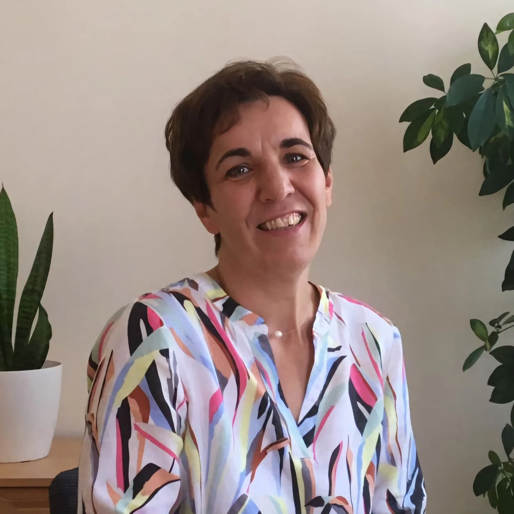 Eine Frau mit kurzem braunem Haar, die auf einem Stuhl sitzt, trägt ein buntes, gestreiftes Hemd und lächelt vor einem hellen Hintergrund mit grünen Zimmerpflanzen.