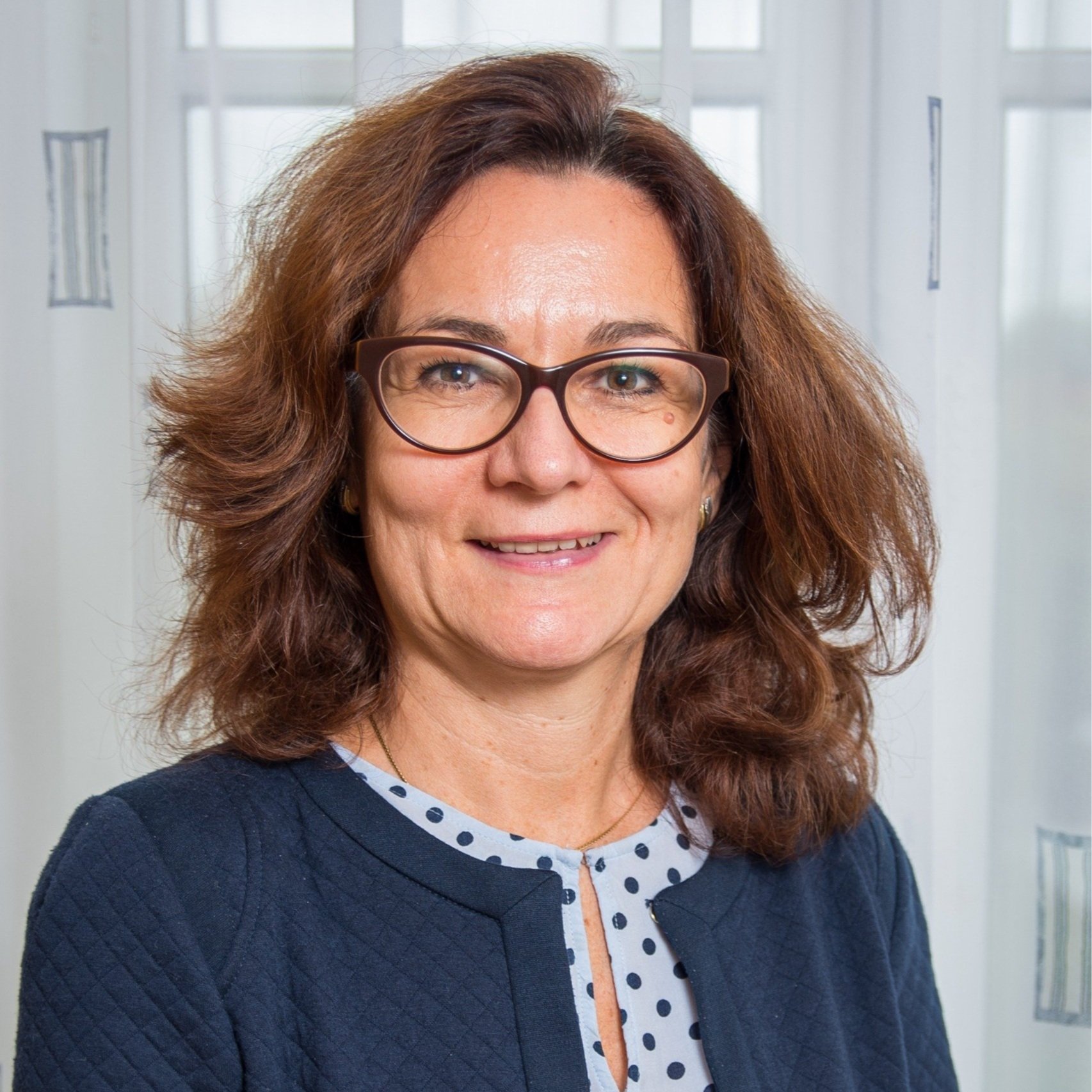 Porträt einer Frau mit braunen, gewellten Haaren und Brille, lächelnd, im Hintergrund ein Bürofenster.