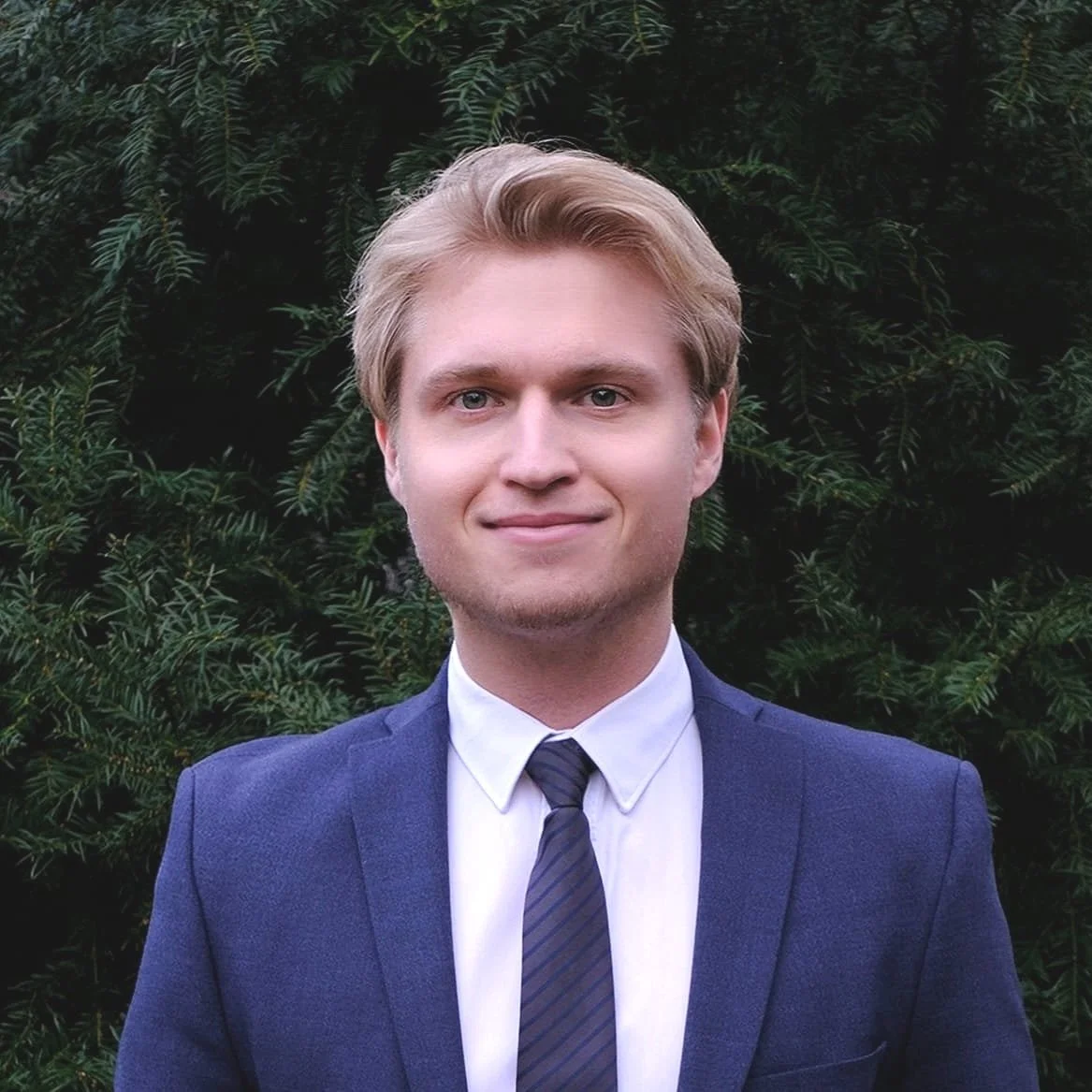 Ein junger Mann in formeller Kleidung vor einem grünen Busch.