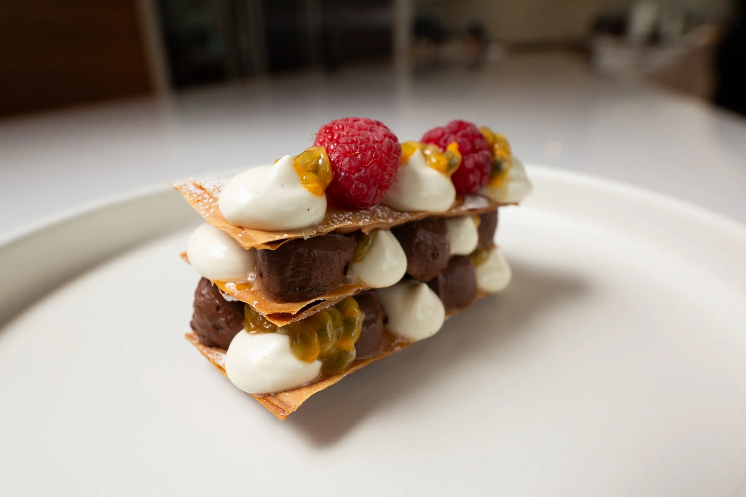 A layered dessert with pastry, whipped cream, chocolate, raspberries, and passion fruit on a white plate.