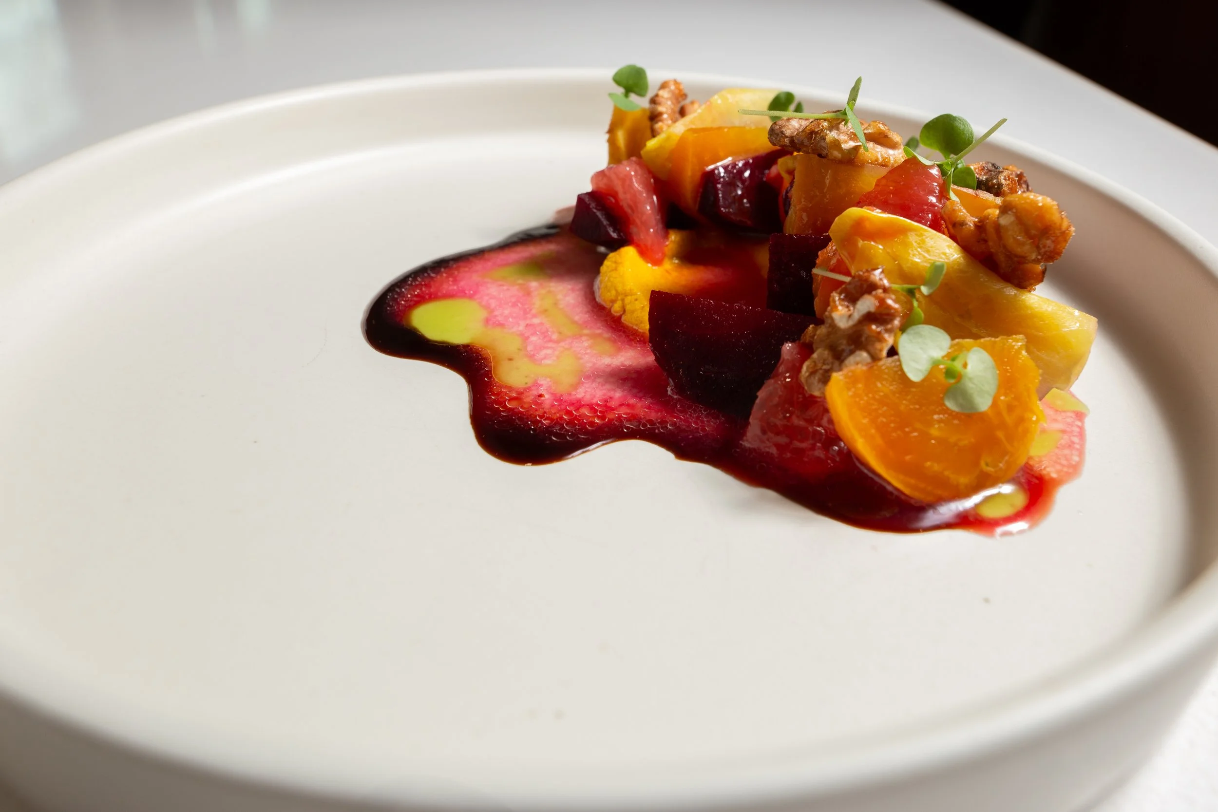 A white plate with a colorful artistic arrangement of sliced beets, carrots, and microgreens, garnished with a sauce drizzle.