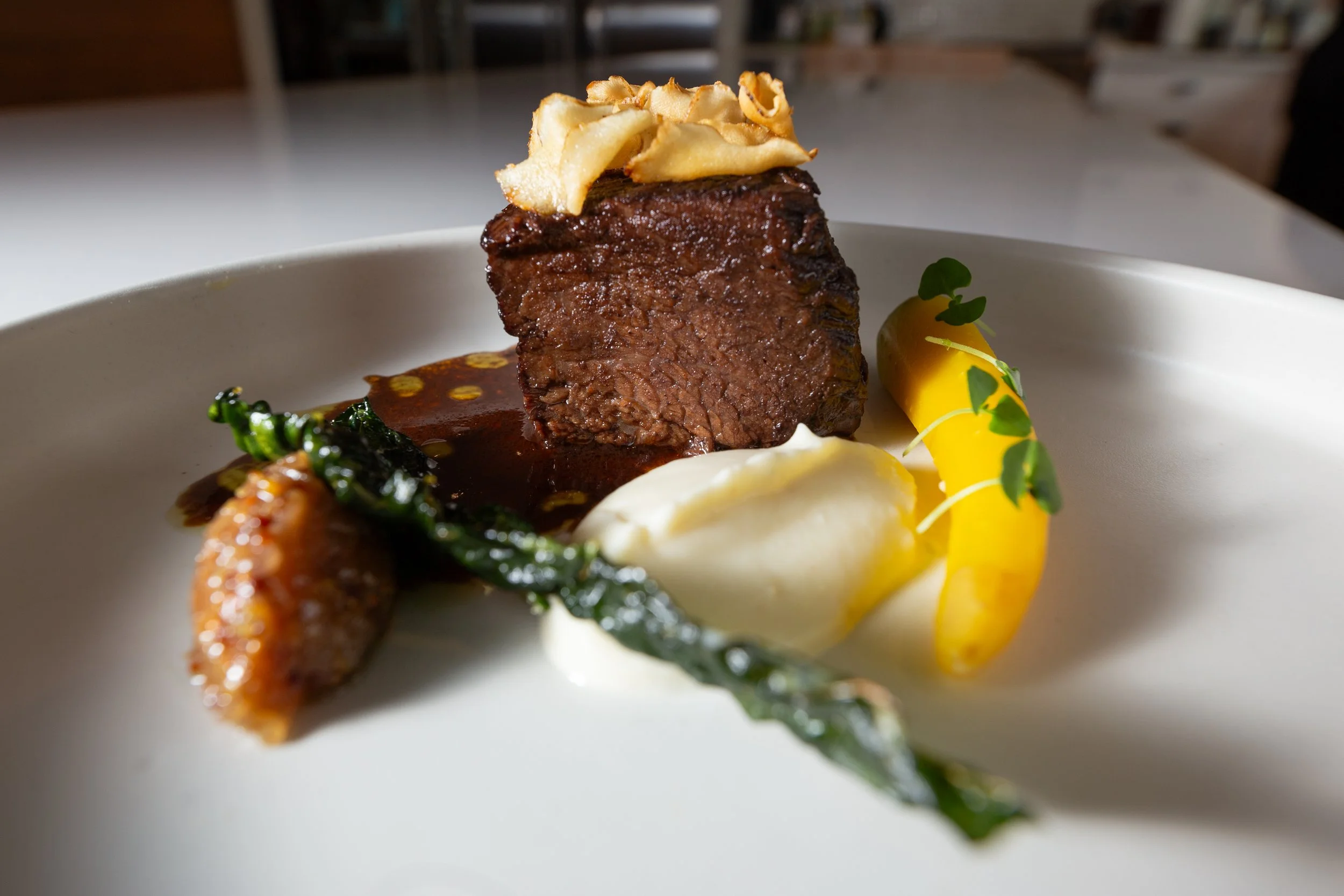 A plated gourmet dish featuring a large cube of braised beef, topped with crispy onion strings, accompanied by a smear of mashed potatoes, a bright yellow pepper, and garnished with microgreens, all on a white plate.