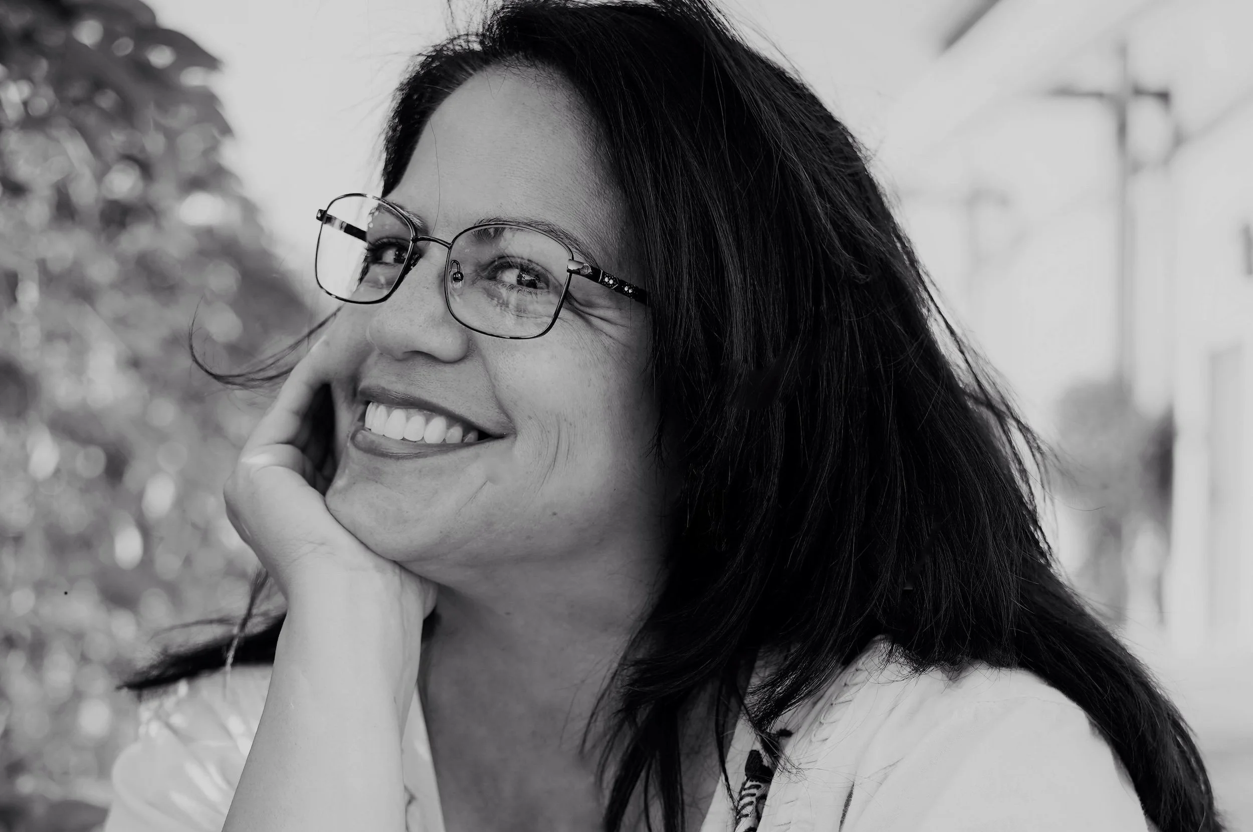 A woman with glasses smiling and resting her chin on her hand outdoors.