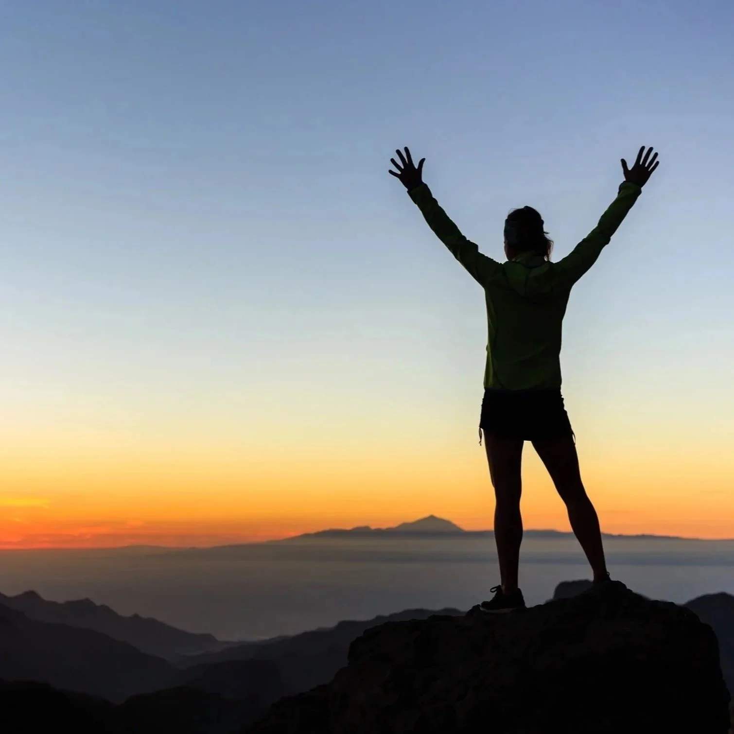 Person standing on a mountain peak with arms raised during sunset, silhouetted against the colorful sky and distant mountains.