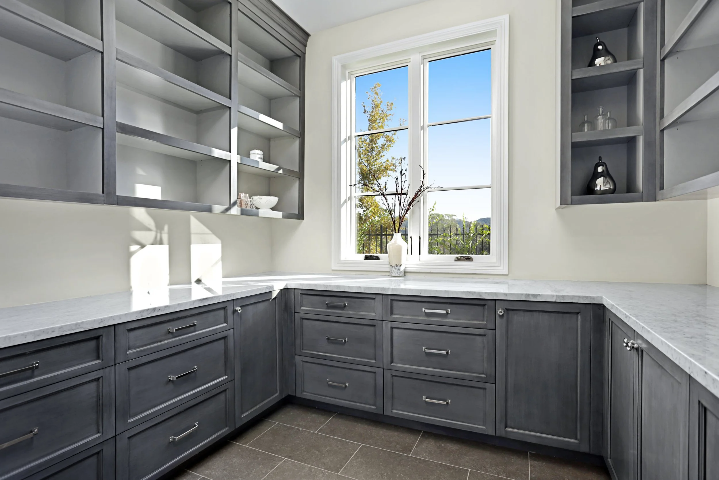 Kitchen corner with gray cabinets, open shelves, marble countertops, and a window with a white frame showing a view of trees and a blue sky.