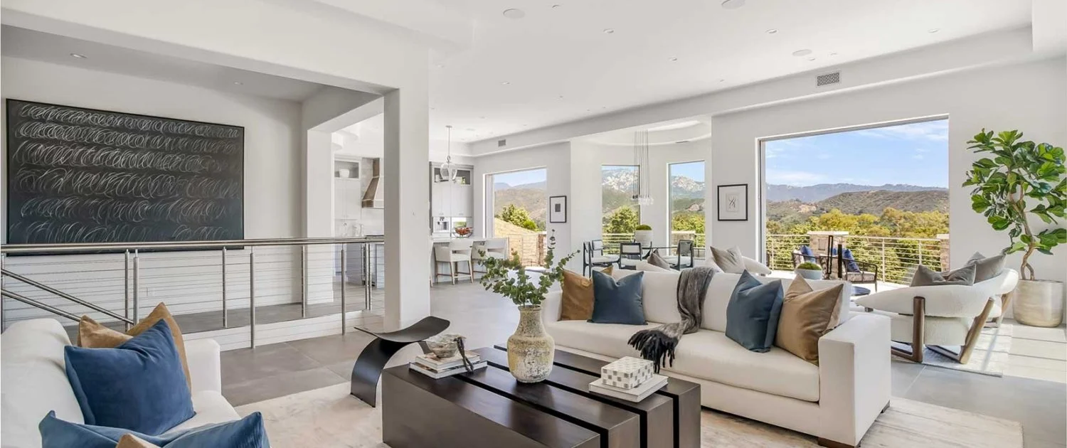 Open living room with white walls, large windows, and mountain views, furnished with white sofas, blue and beige pillows, a black coffee table with a decorative vase and books, and a white armchair with a small table, in a modern house.