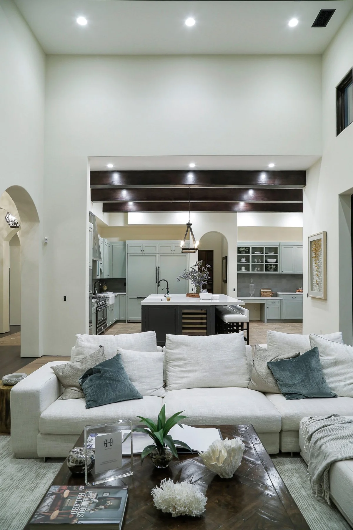 Living room with a white sofa and dark throw pillows, wooden coffee table with decorations, and an open kitchen with an island and cabinetry in the background.