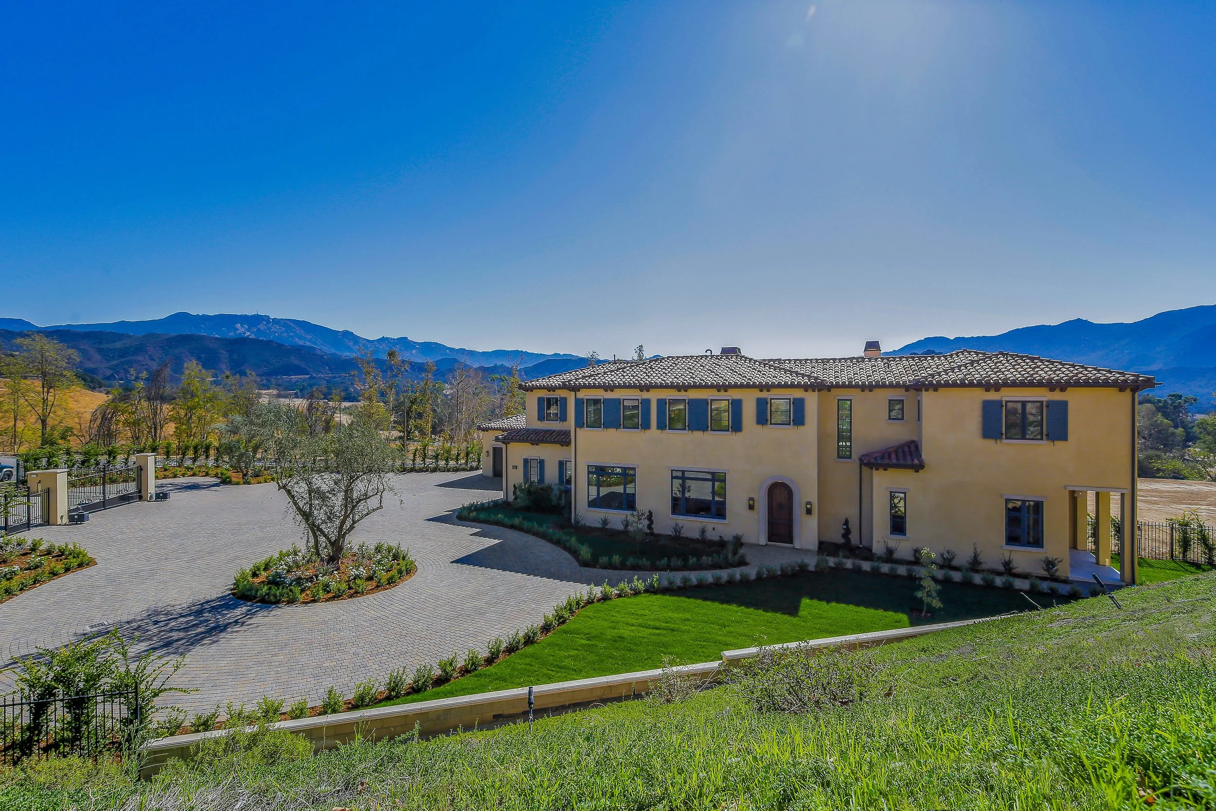 A large two-story house with yellow walls and a terracotta tile roof, surrounded by landscaped gardens and a paved driveway, with mountains in the background under a clear blue sky.