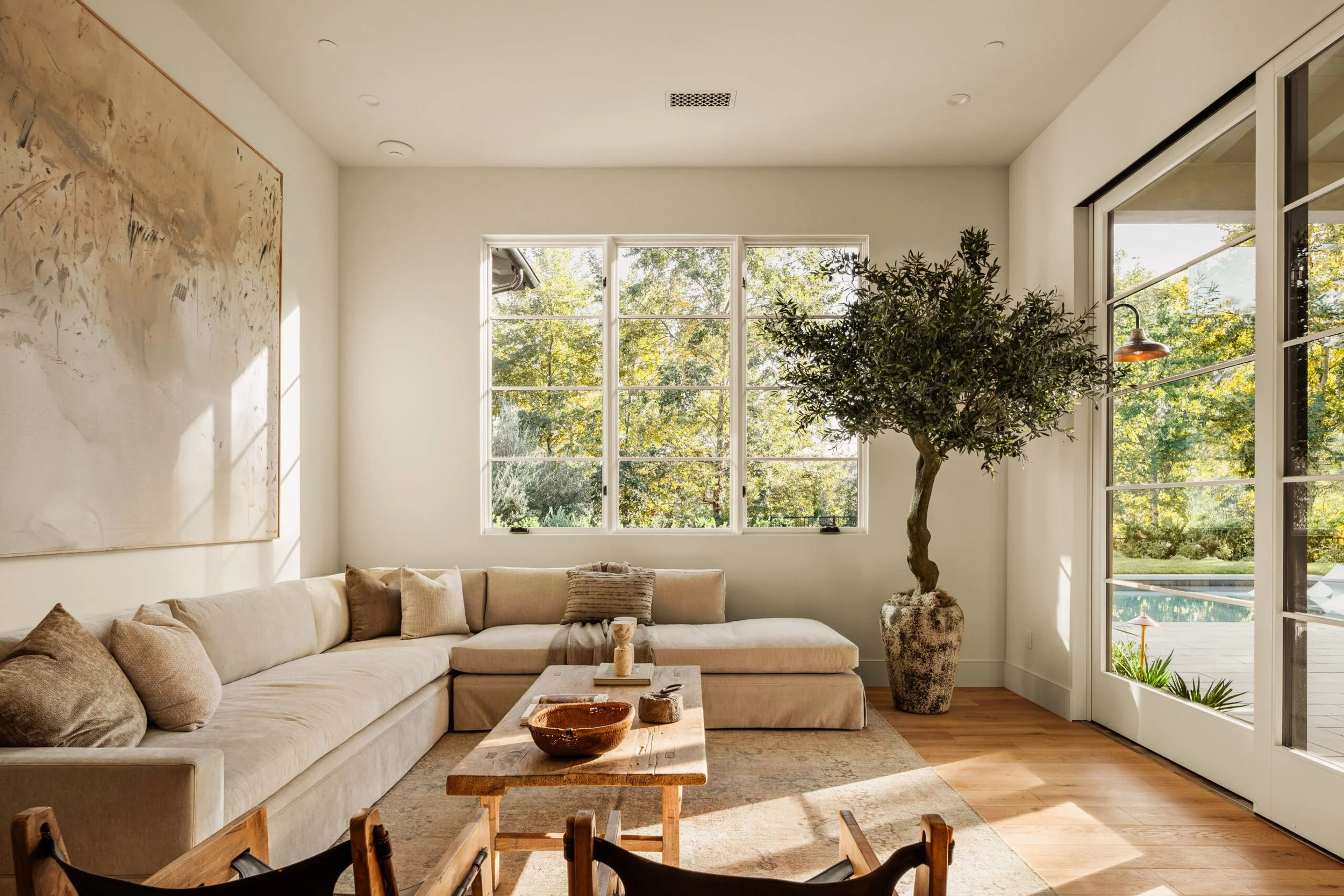 Living room with beige sectional sofa, a wooden coffee table, a large abstract painting on the wall, a potted tree, and windows showing a backyard with greenery and a pool.