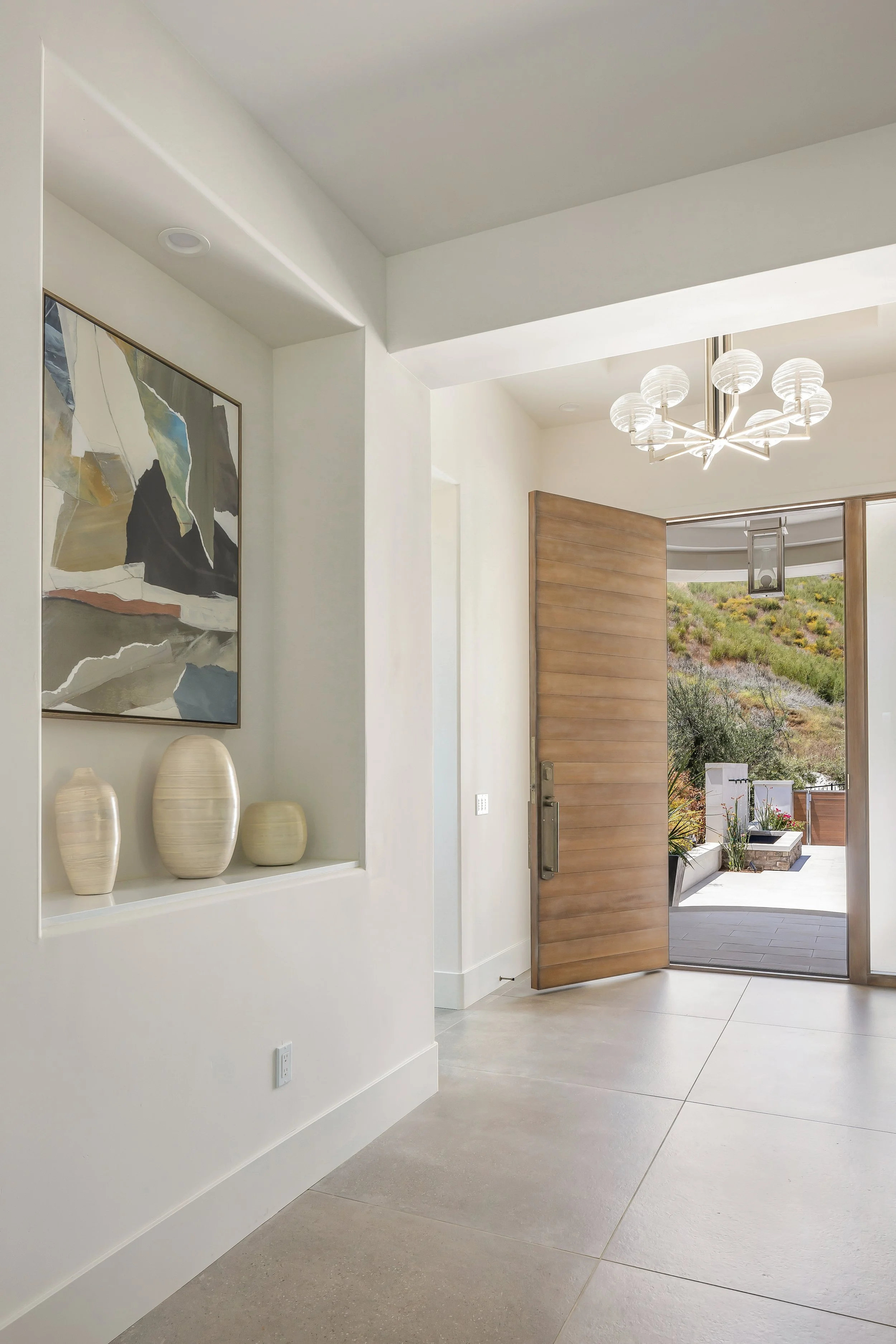 Modern home entryway with open wooden door, white walls, abstract painting, decorative vases, and outdoor patio with plants and hillside view.