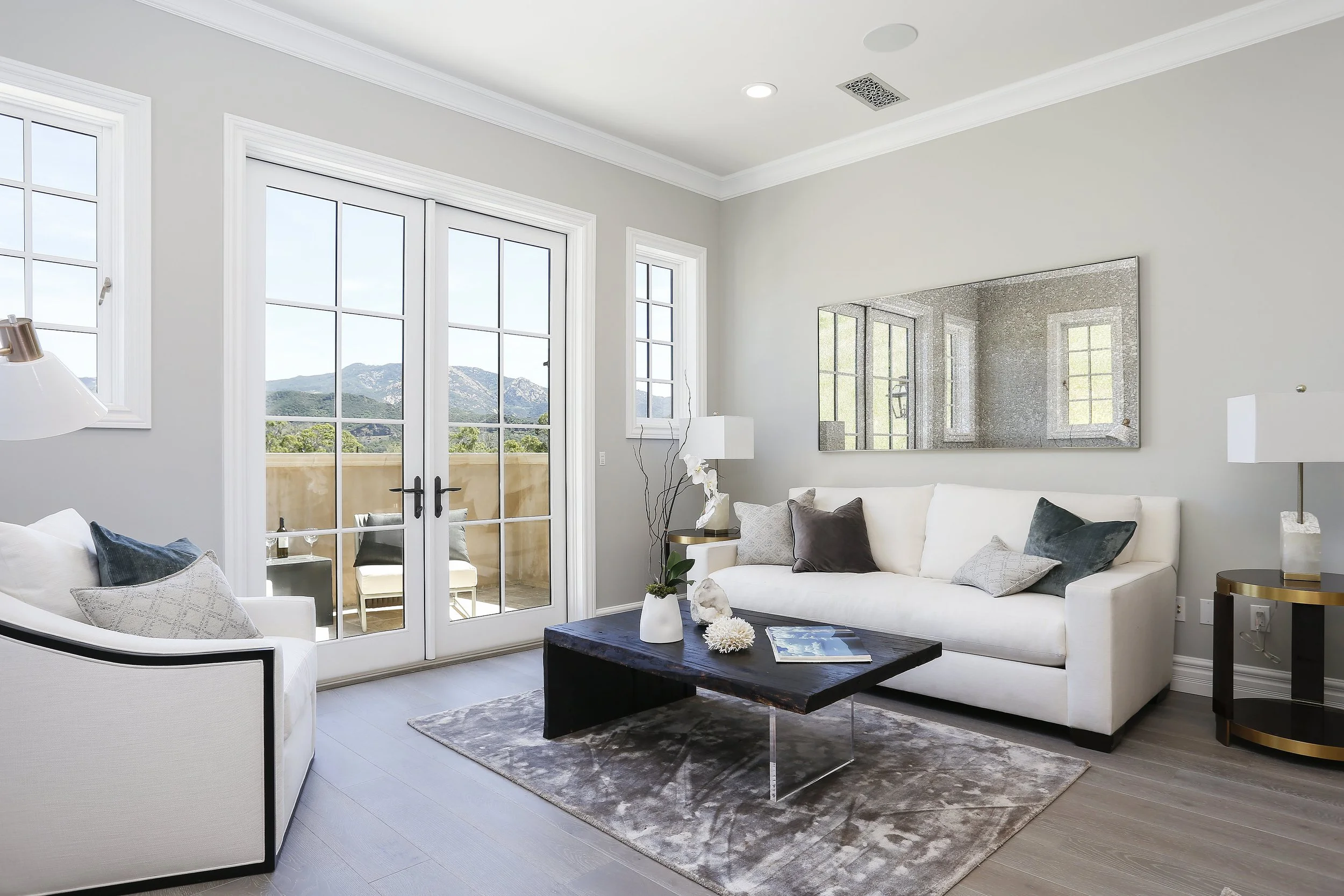 Bright living room with a white sofa, glass double doors leading to a balcony, mountain landscape view, modern decor, and a large mirror on the wall.