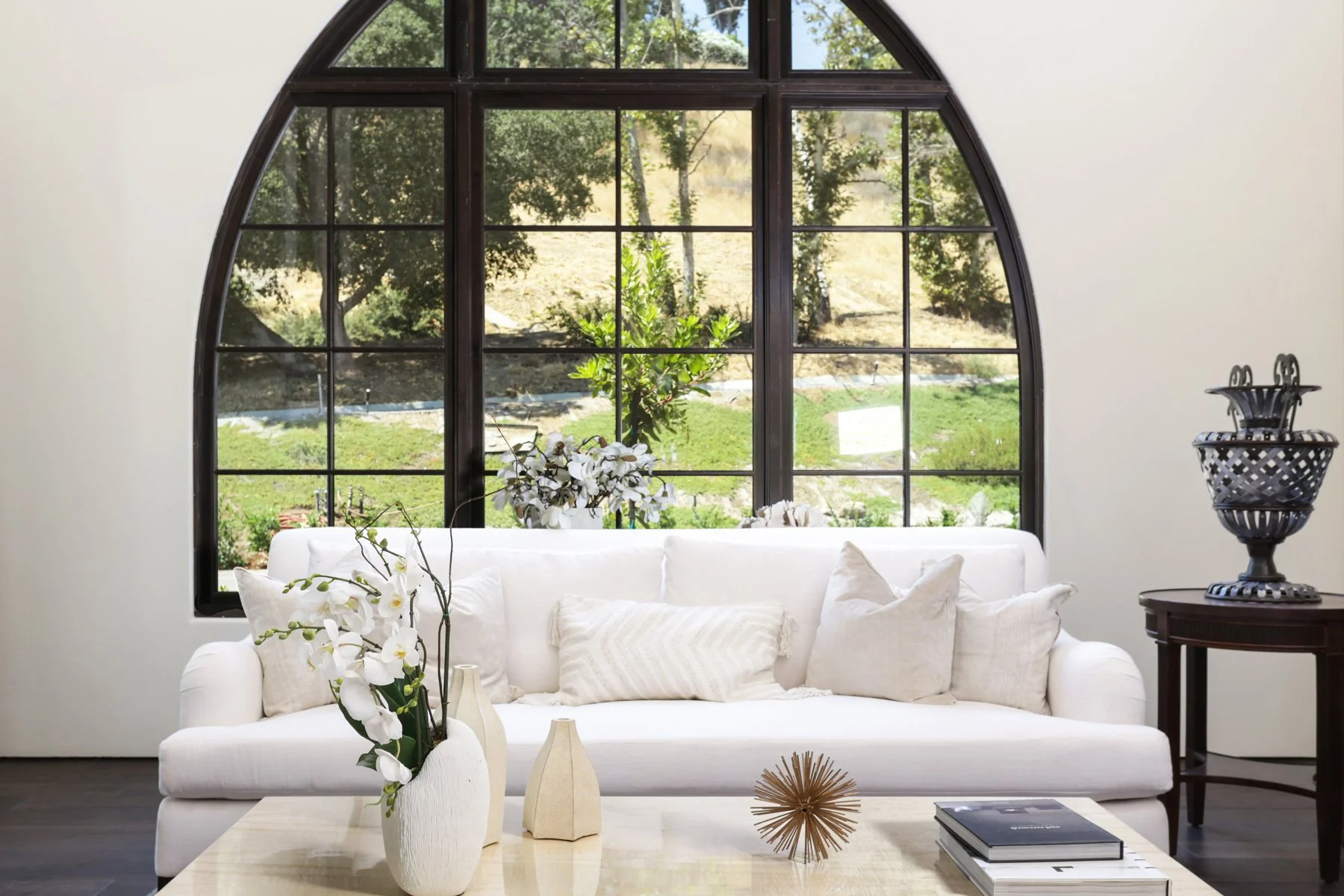 Living room with a large arched window, white sofa with pillows, and decorative vases and objects on a coffee table, with outdoor greenery visible through the window.