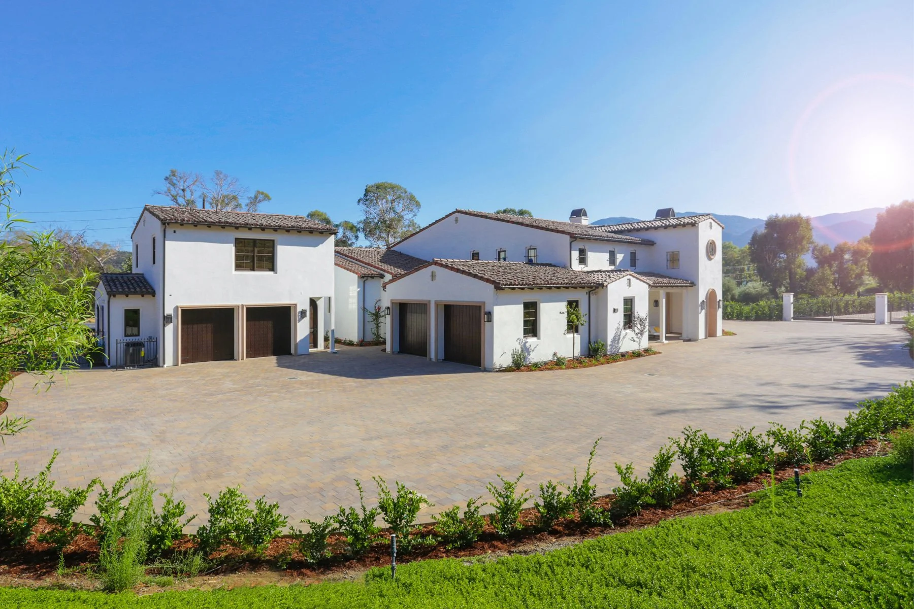 A large white Mediterranean-style house with multiple levels, red tile roof, and dark brown garage doors, surrounded by manicured green lawns and shrubbery, under a clear blue sky with the sun shining.