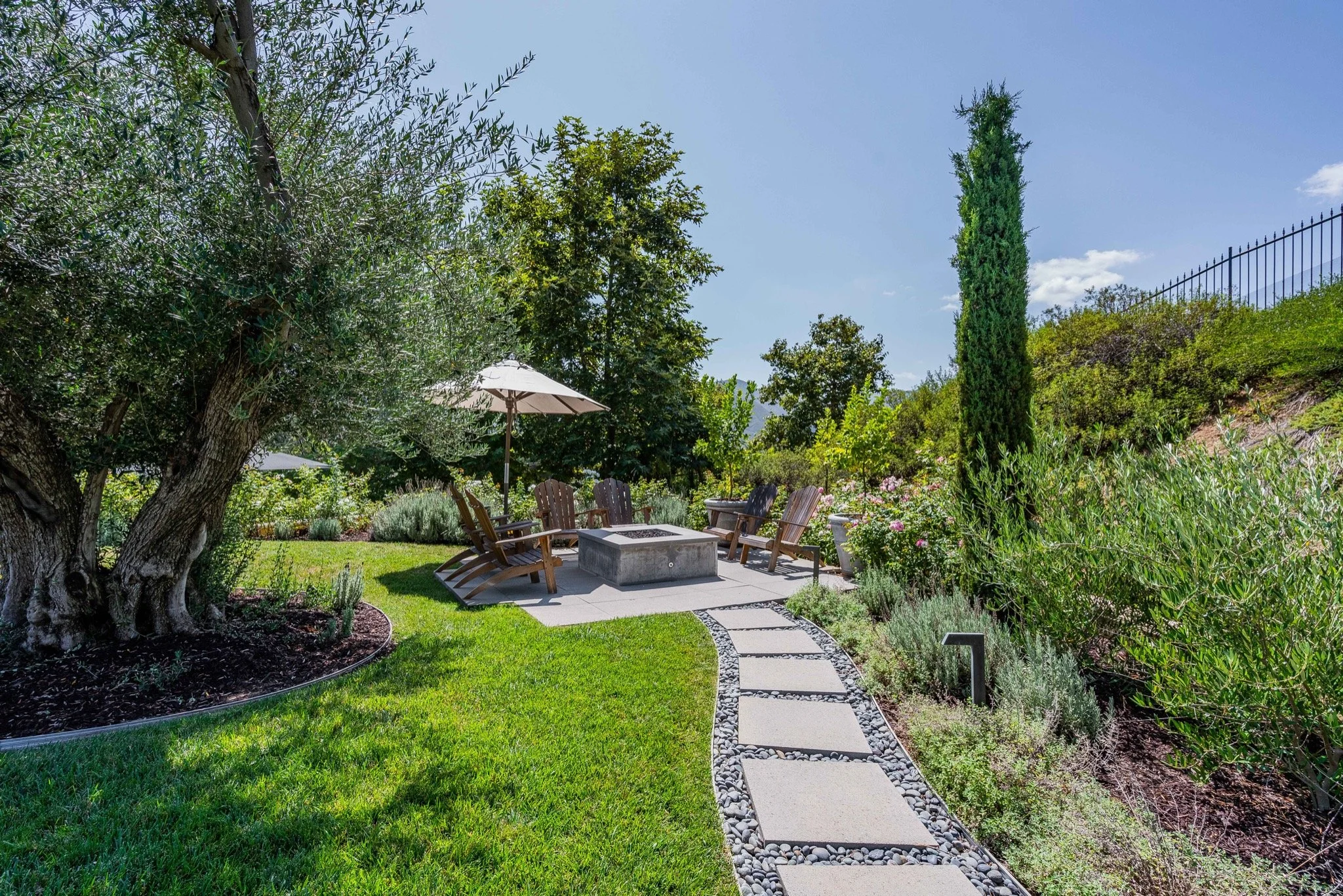 A garden patio with a circular fire pit, surrounded by wooden Adirondack chairs and a white patio umbrella. A stone walkway leads to the seating area, with lush green grass and assorted plants and trees around.