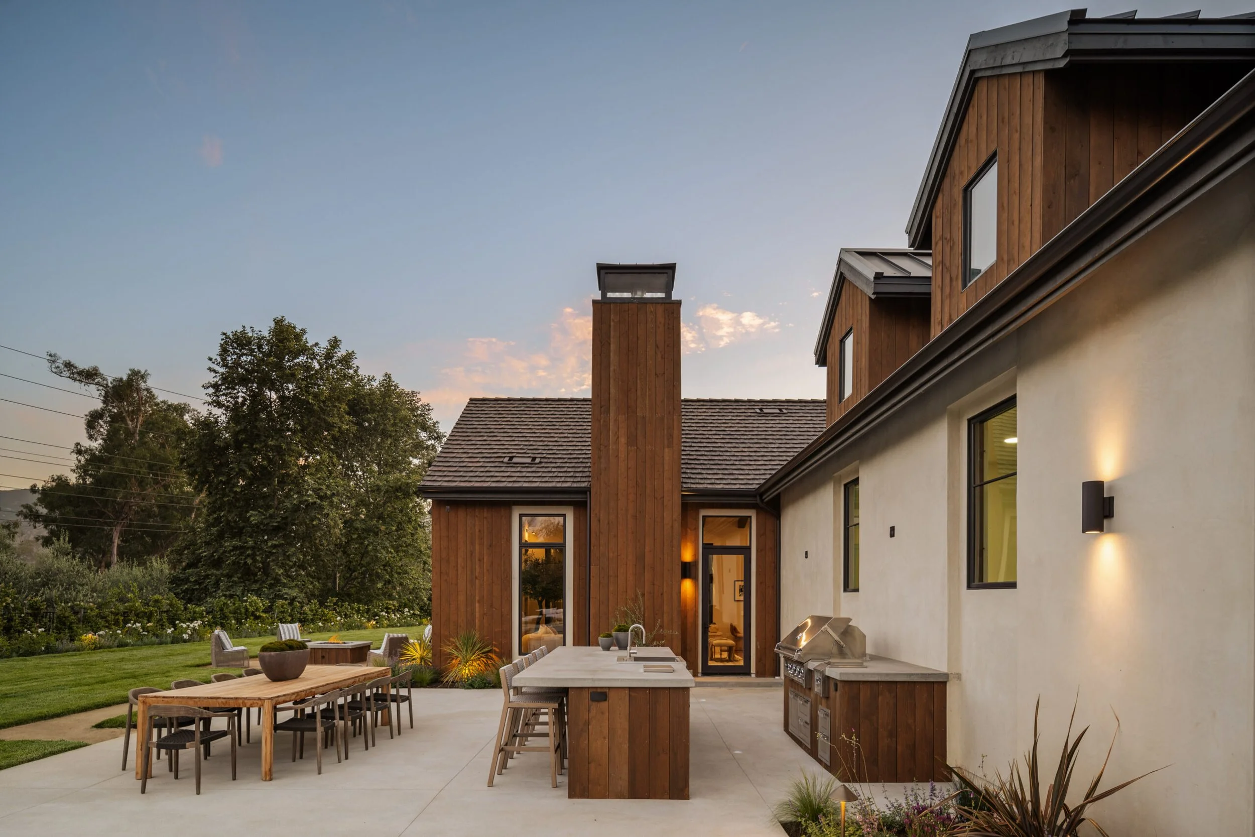 Modern house exterior with outdoor kitchen and dining area at sunset, featuring a wooden and stucco facade with large windows, outdoor grill, and patio furniture, surrounded by greenery