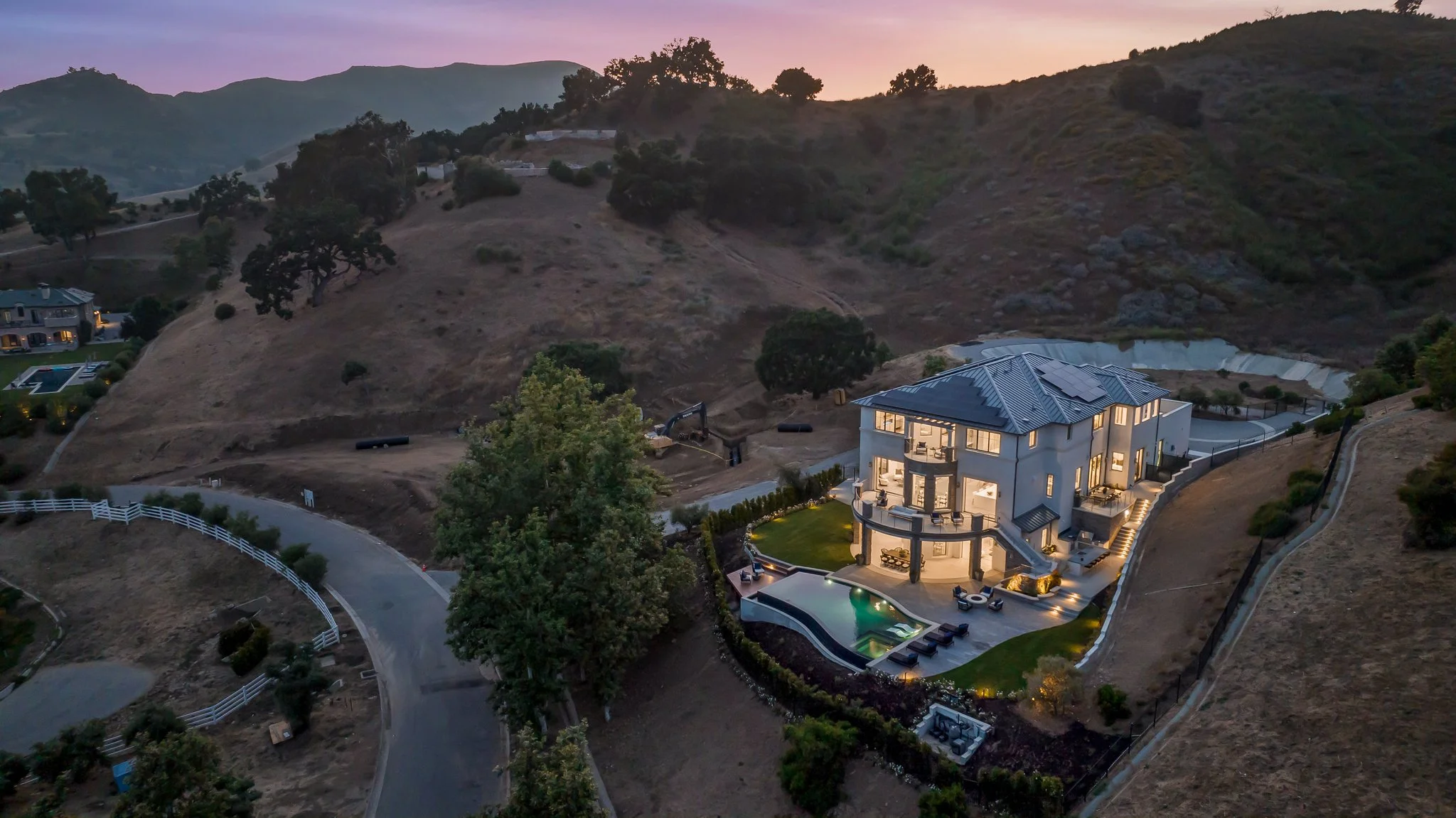 A large, modern multi-story house with outdoor lighting, swimming pool, and landscaped yard, situated on a hillside with hills and trees in the background during dusk.