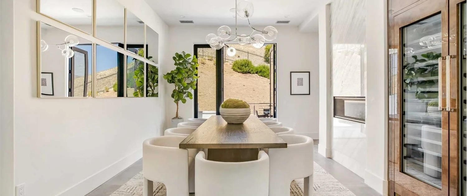 Modern dining room with white chairs around a wooden table, large window doors, and a glass cabinet on the right, with a view of an outdoor hillside.