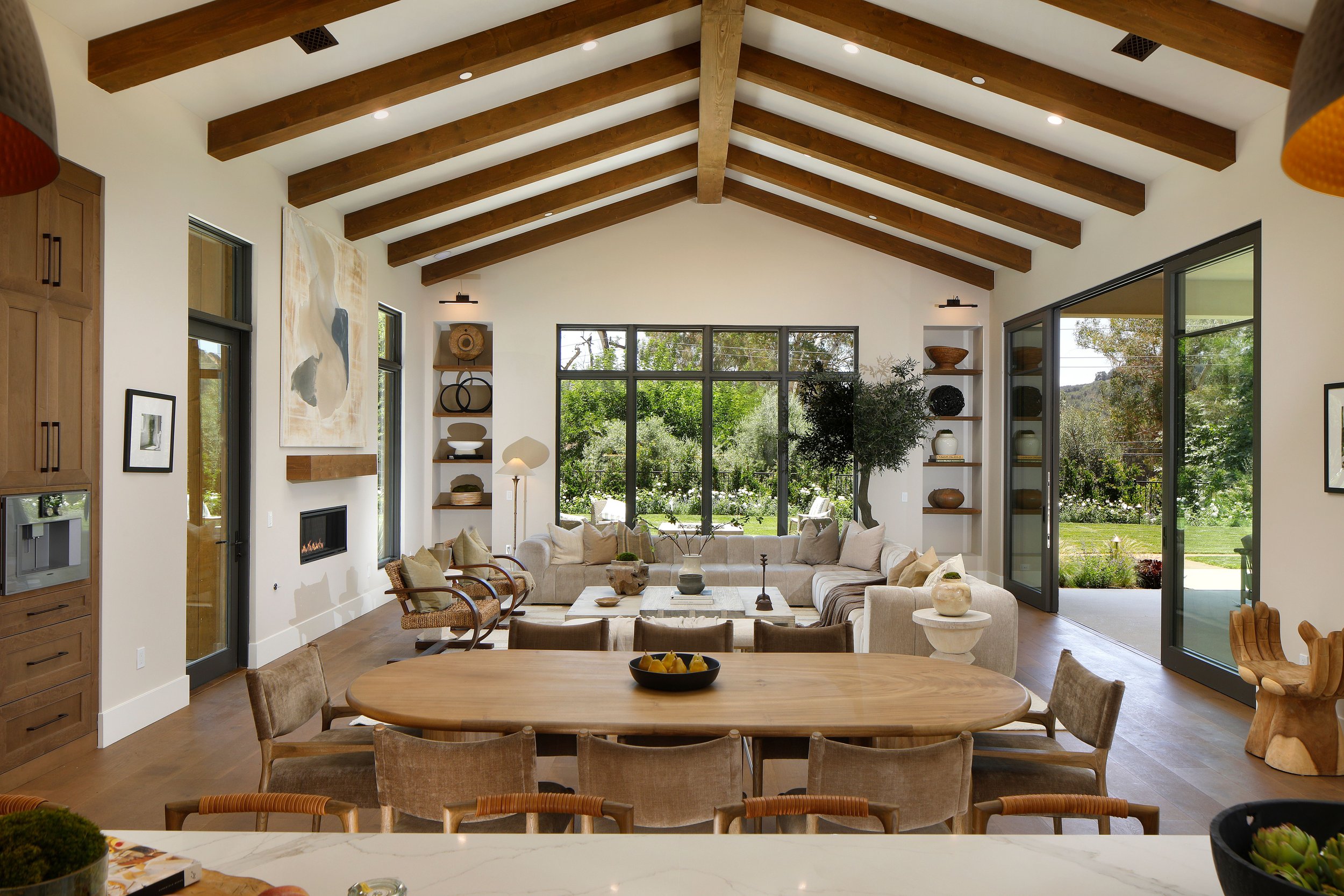 Spacious living room with high vaulted ceiling featuring wooden beams, large windows and sliding glass doors, beige sectional sofa, armchairs, wooden dining table with chairs, decorative shelves with vases, and a view of a lush green garden.