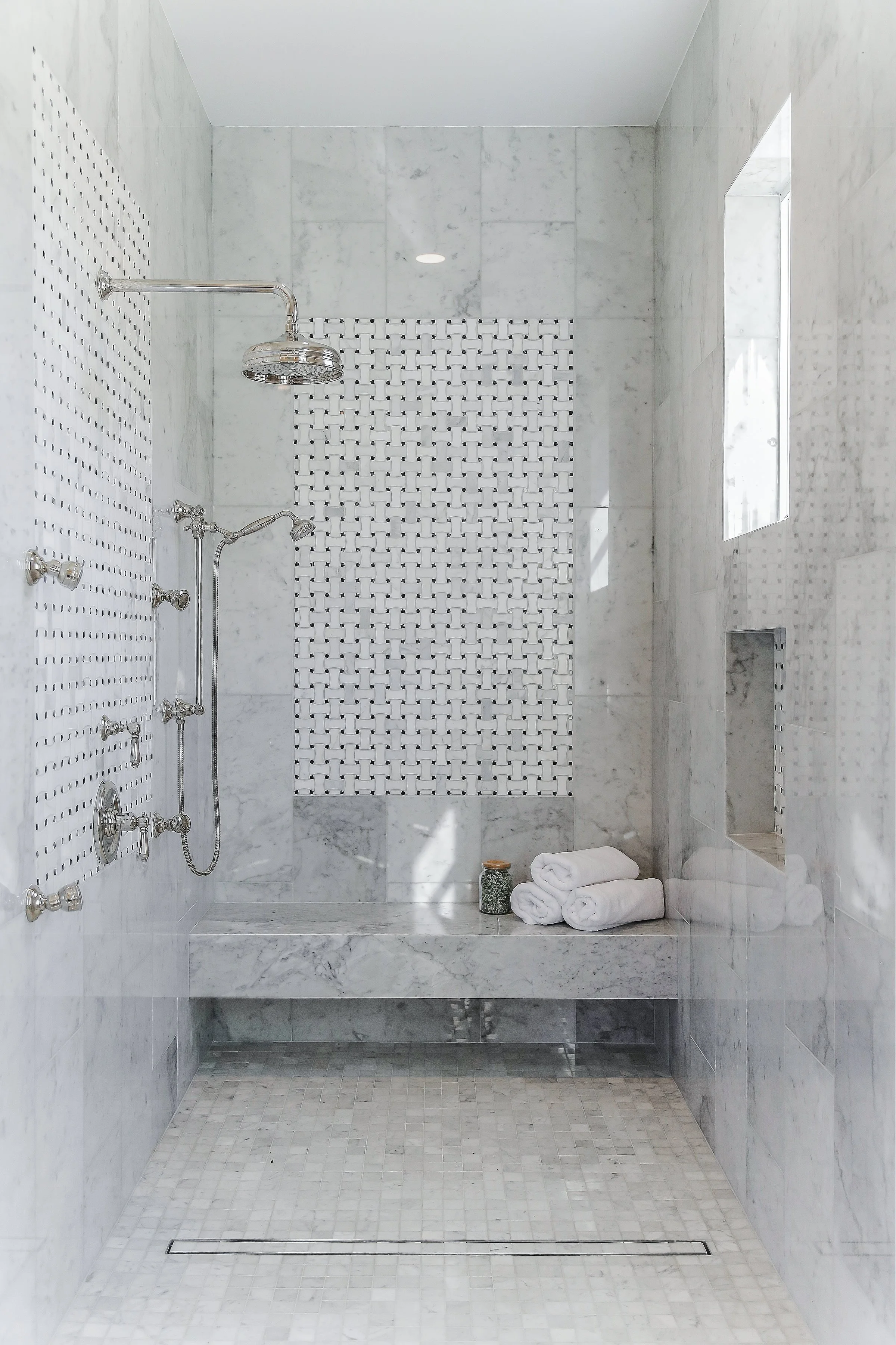 Modern marble shower with built-in niche, multiple shower heads, and rolled white towels on a marble bench