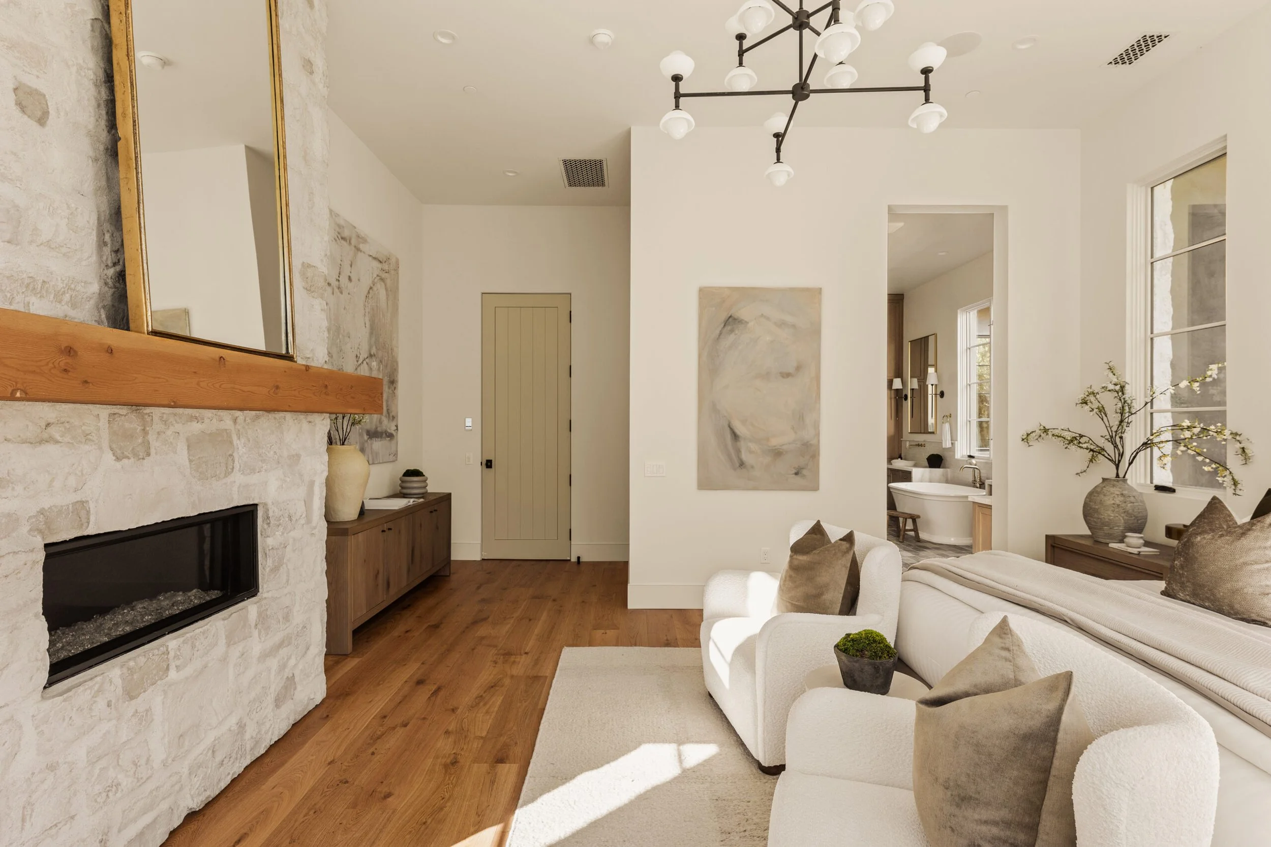 A living room with a white sofa, beige and brown pillows, a wooden side table with a large vase and decorative items, a stone wall with a fireplace, a large mirror, a modern chandelier, a painting, and a spacious layout with a bathroom visible throug