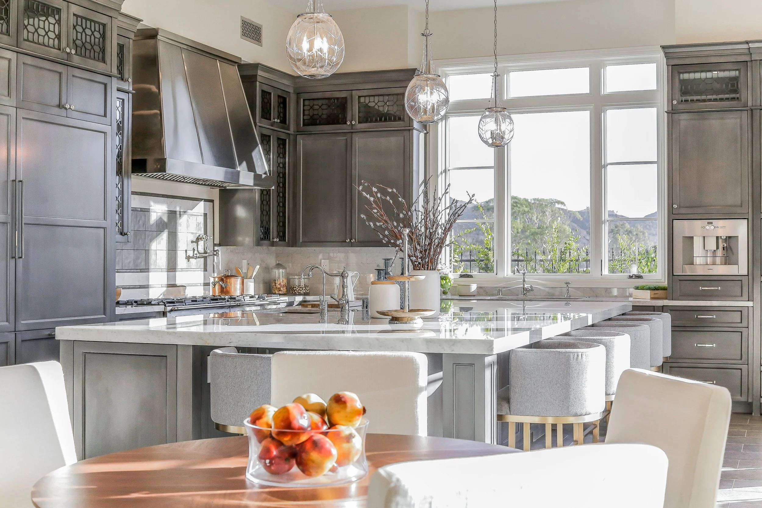 Modern kitchen with gray cabinets, stainless steel range hood, white marble countertops, large window with a view of trees, and hanging pendant lights. A bowl of apples on the dining table.