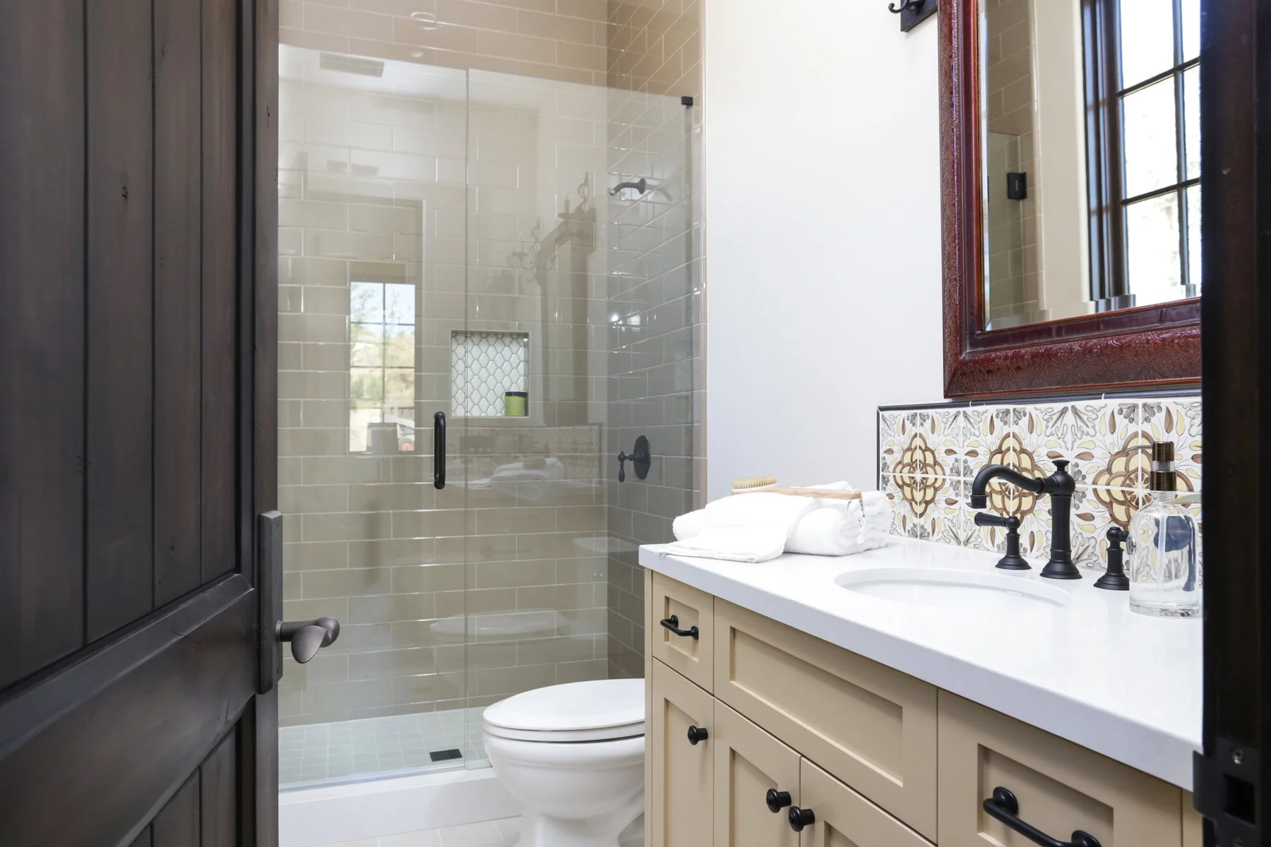 A modern bathroom with a walk-in glass shower, beige tiled walls, a white toilet, a cream-colored vanity with a white countertop, a black faucet, and a large wooden-framed mirror. There are decorative tiles behind the sink and a window in the shower 