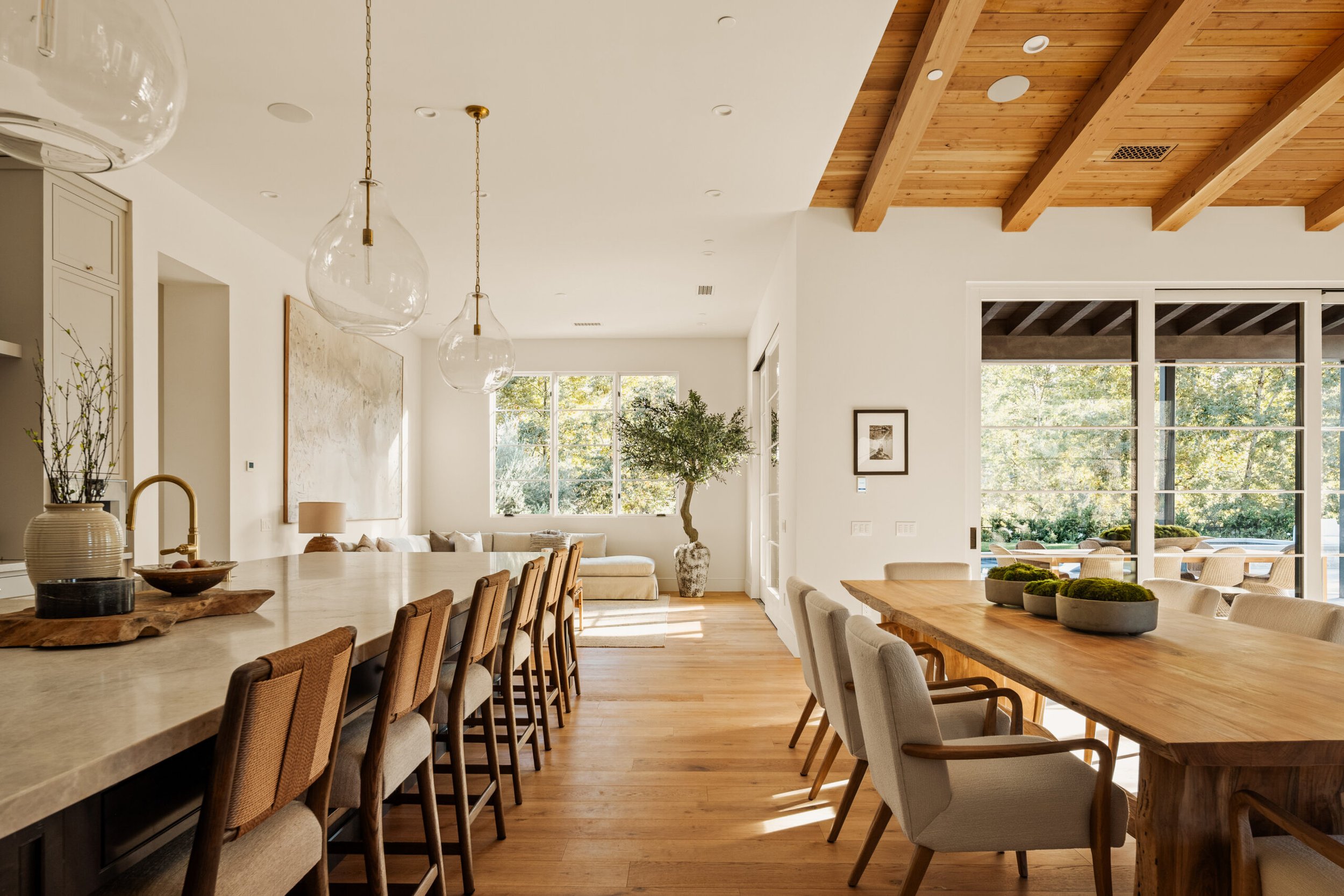 Bright, modern open-concept dining and living area with wooden flooring, large windows, a wooden dining table with gray chairs, and a kitchen island with barstools.