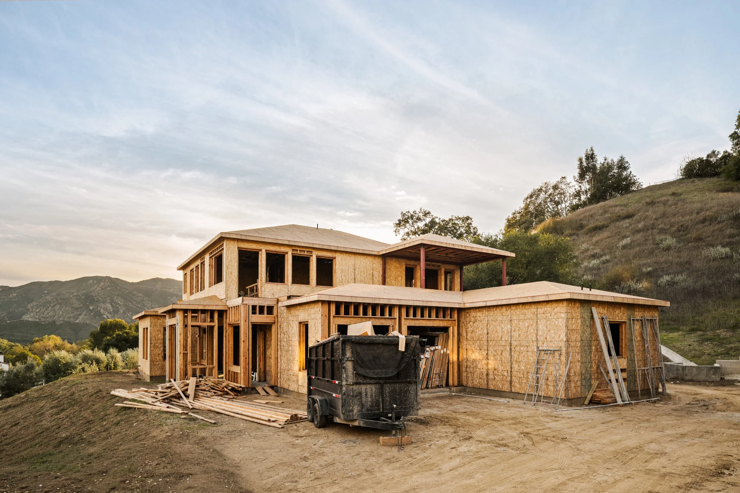 Under-construction house with wooden framing, construction equipment and materials on dirt ground, hillside with trees and mountains in the background.