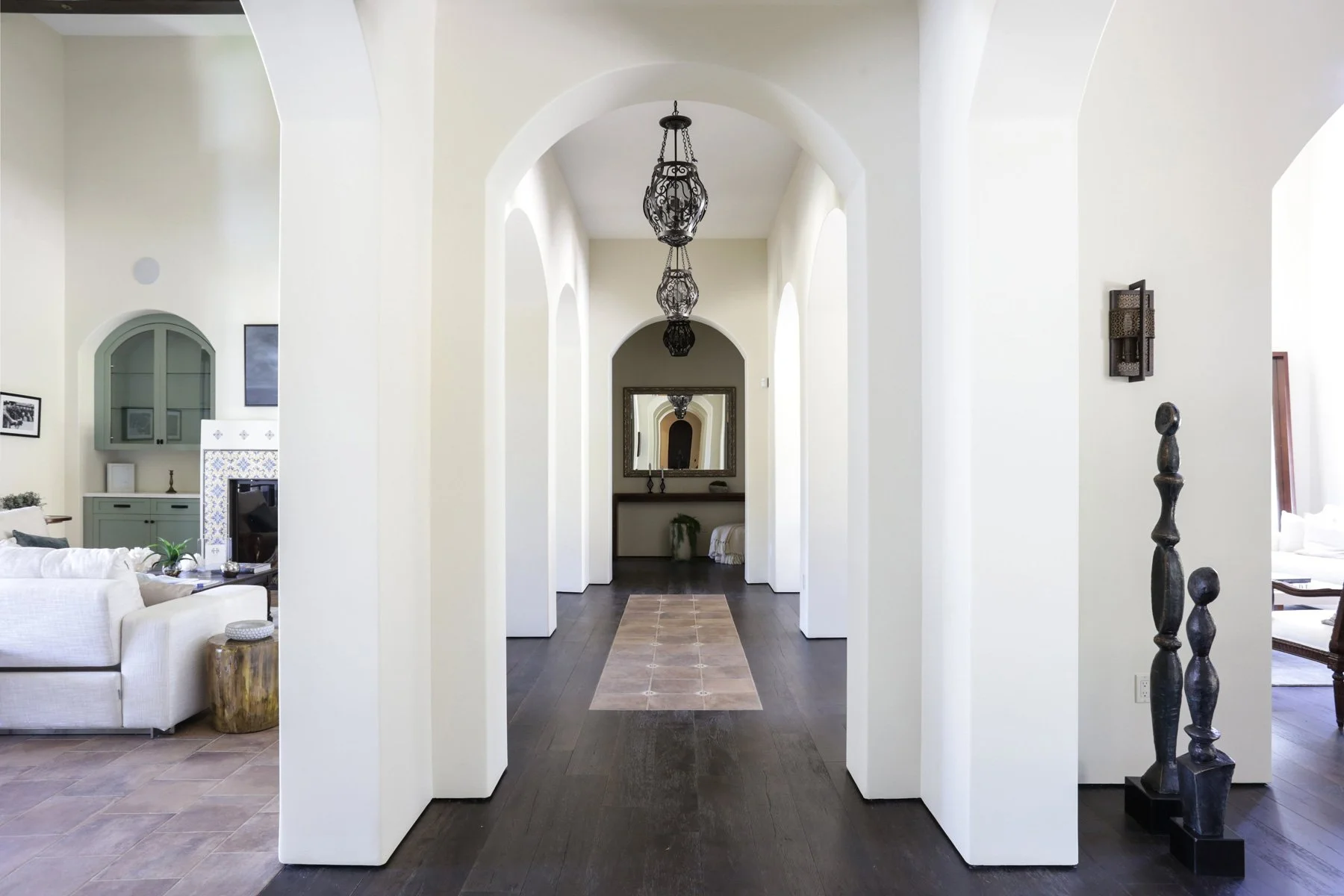 Interior view of a home with a hallway featuring arched openings, dark wooden floors, hanging lantern-style lights, and a mirror at the end of the hall. Part of a living room with white and green furniture and decorative items is visible on the left,