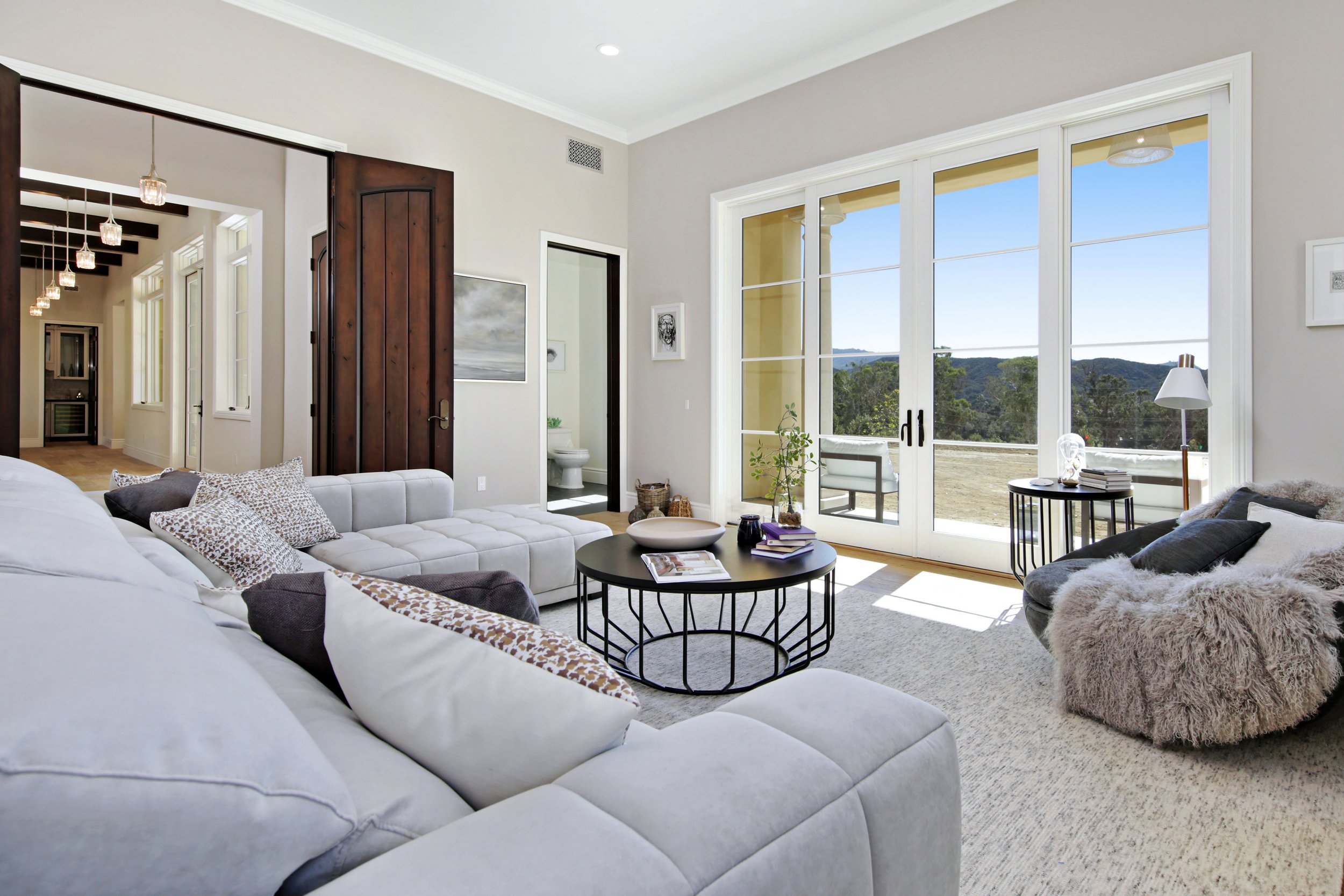 Living room with large windows overlooking scenic outdoor view, white sectional sofa with patterned pillows, black round coffee table, cozy armchair with throw blanket, side table with lamp, open door to bathroom, and decorative artwork on walls.