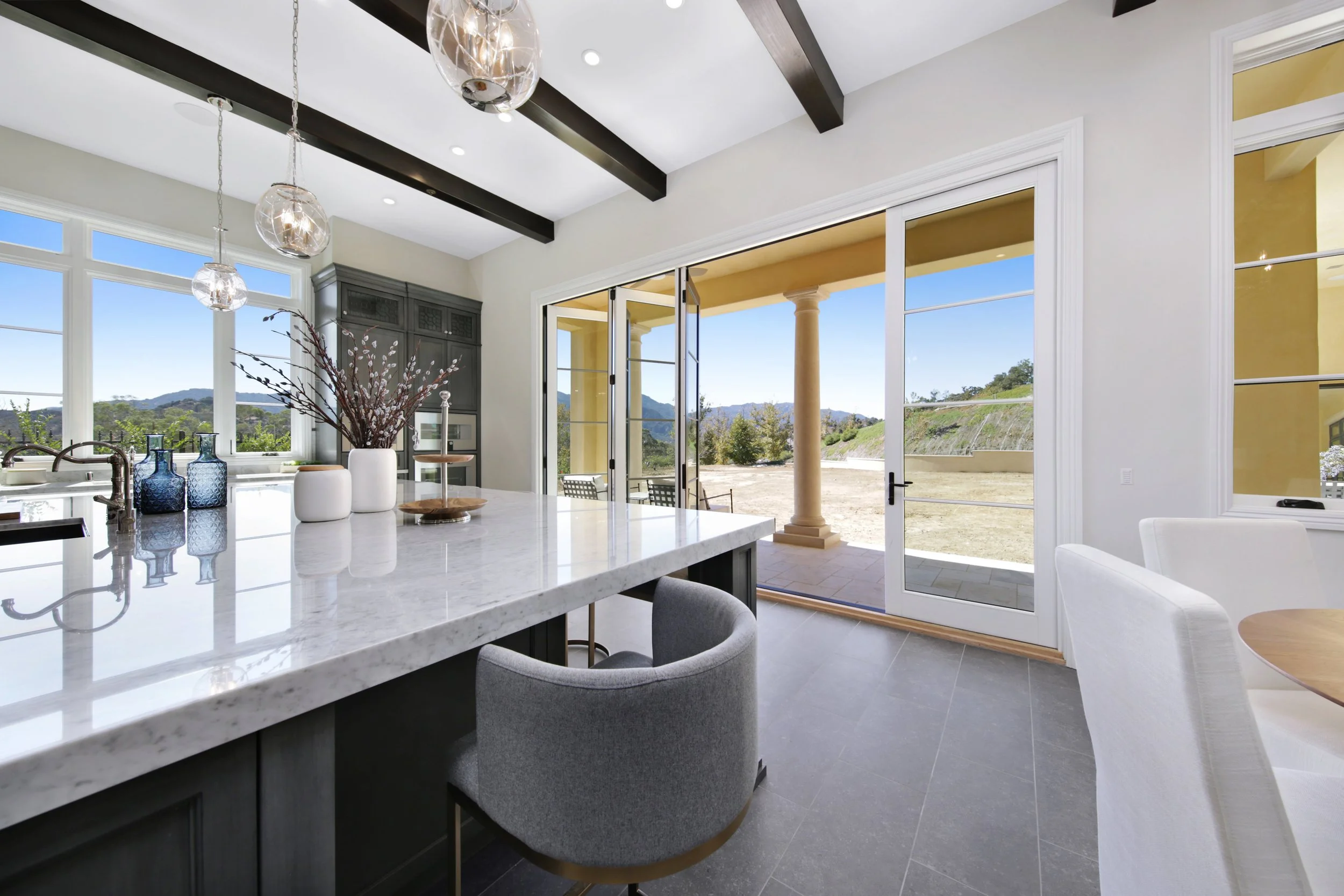 Modern kitchen with white marble island, gray chairs, and large glass sliding doors opening to outdoor patio with columns and mountainous landscape.