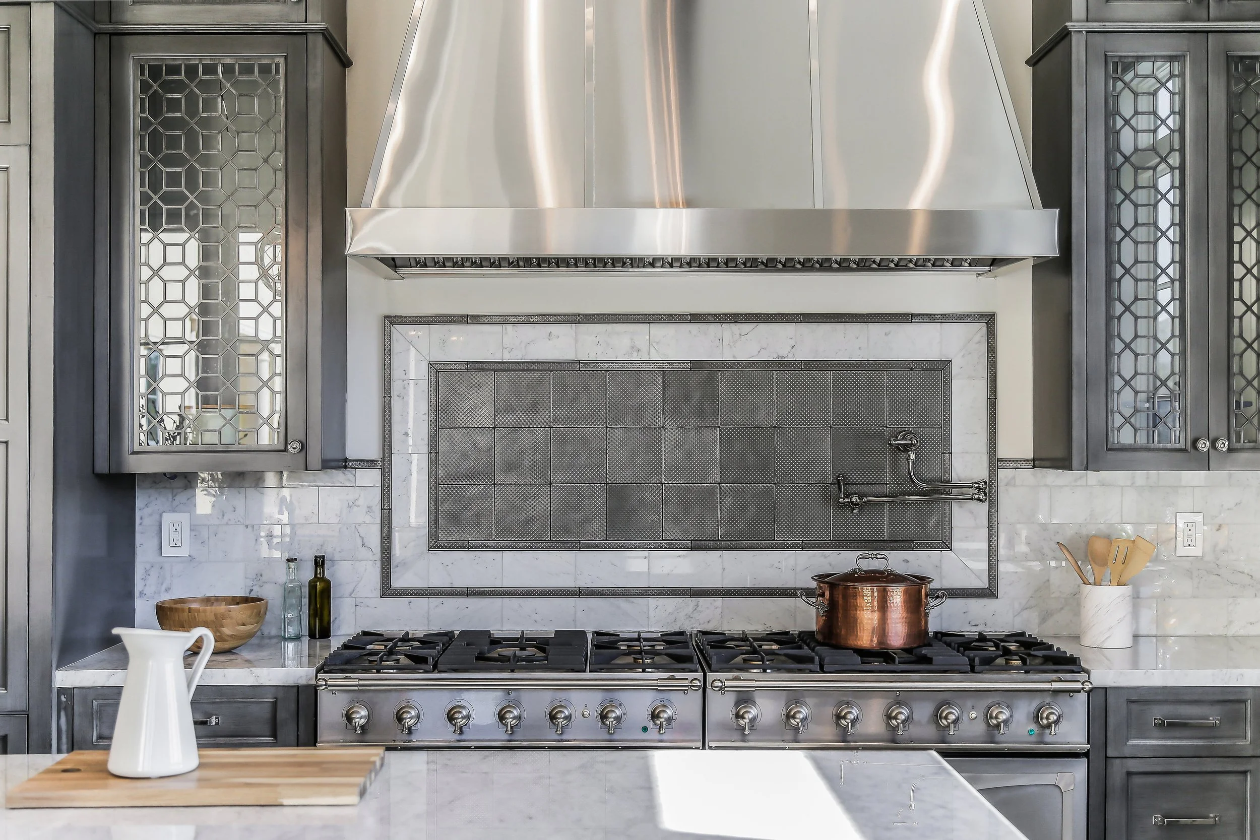 Modern kitchen with stainless steel range hood, six-burner stove, copper pot, marble backsplash, and gray cabinetry.