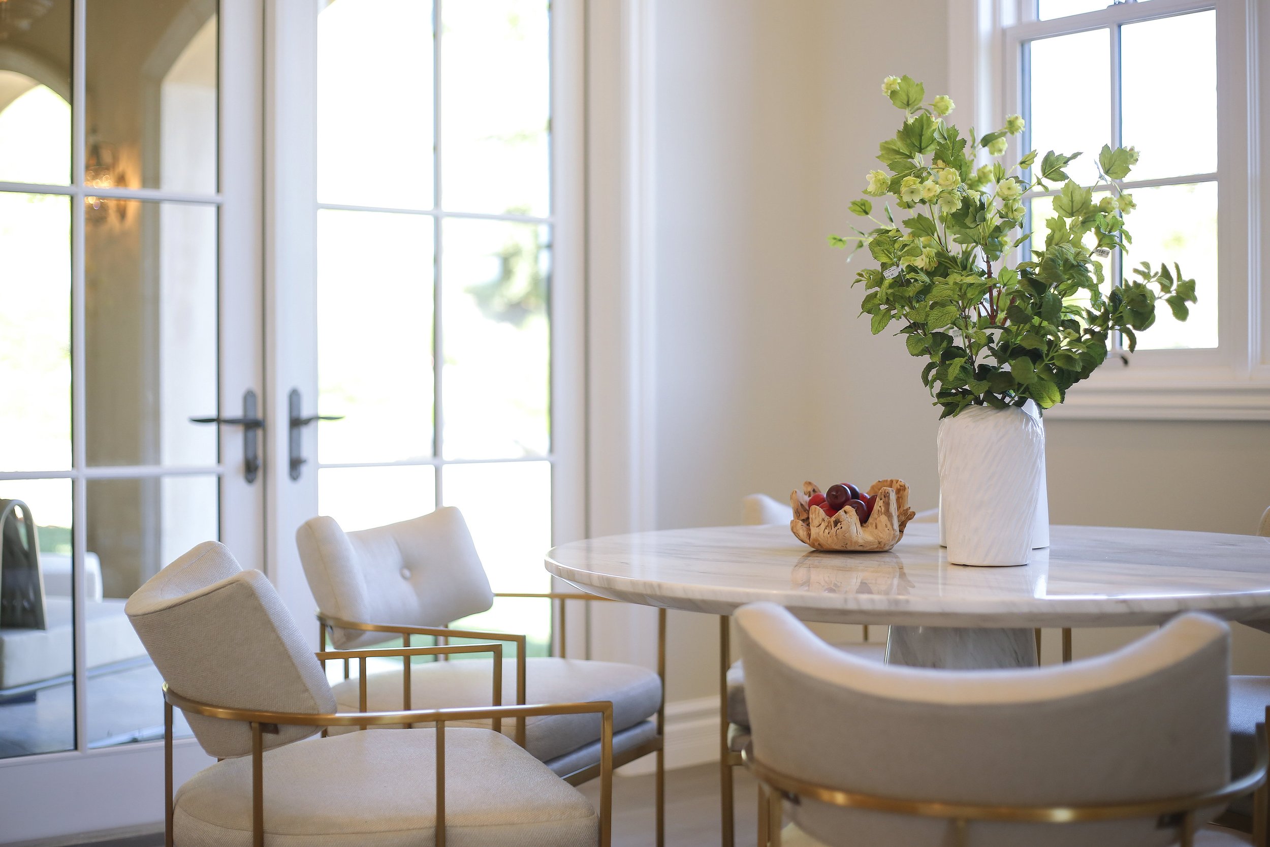 Dining room with a white marble table, a large vase with green leaves, a bowl of cherries, and beige chairs with golden armrests, illuminated by natural light from windows and glass doors.