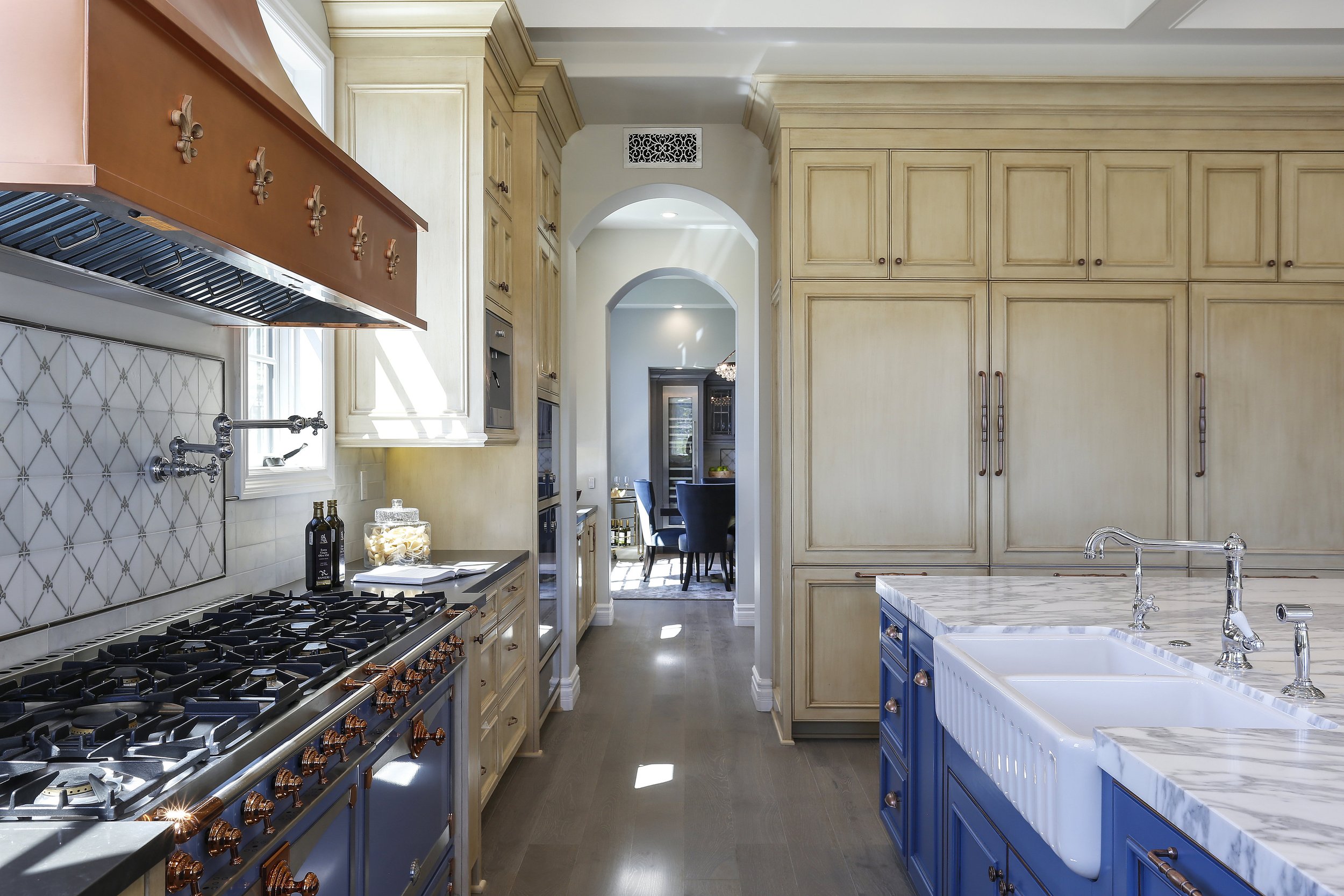 Spacious kitchen with cream cabinets, a blue island with a marble countertop, and a farmhouse sink. Sunlight streams through a window, illuminating the stove with copper hardware and a patterned backsplash. In the background, a dining area is visible