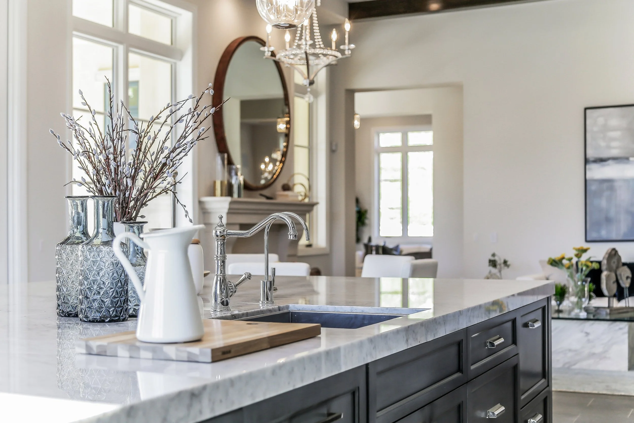 Modern kitchen with black cabinetry, white marble countertop, and decorative vases, with a view into a bright living space with large windows.