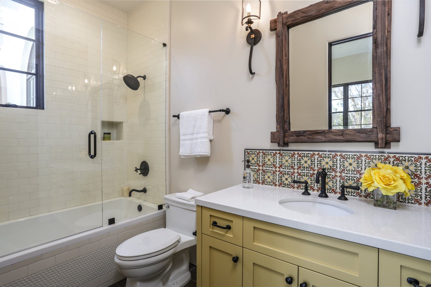 Bathroom with a glass-enclosed shower, a white toilet, a yellow vanity with black hardware, a white countertop with a sink, a mirror with a rustic wooden frame, a yellow flower arrangement, and decorative backsplash tiles, with a window letting in na