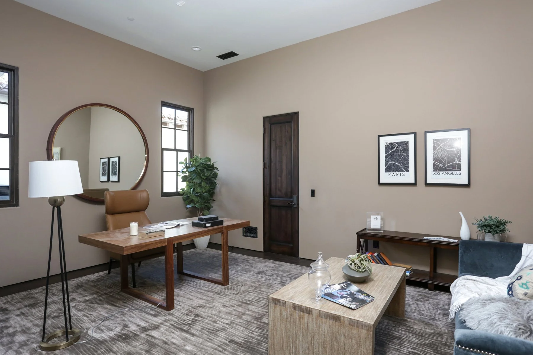 Modern office with beige walls, wooden desk, tan leather chair, large round mirror, potted plant, framed black-and-white city map prints, a wooden side table, and a blue sofa with a throw blanket.