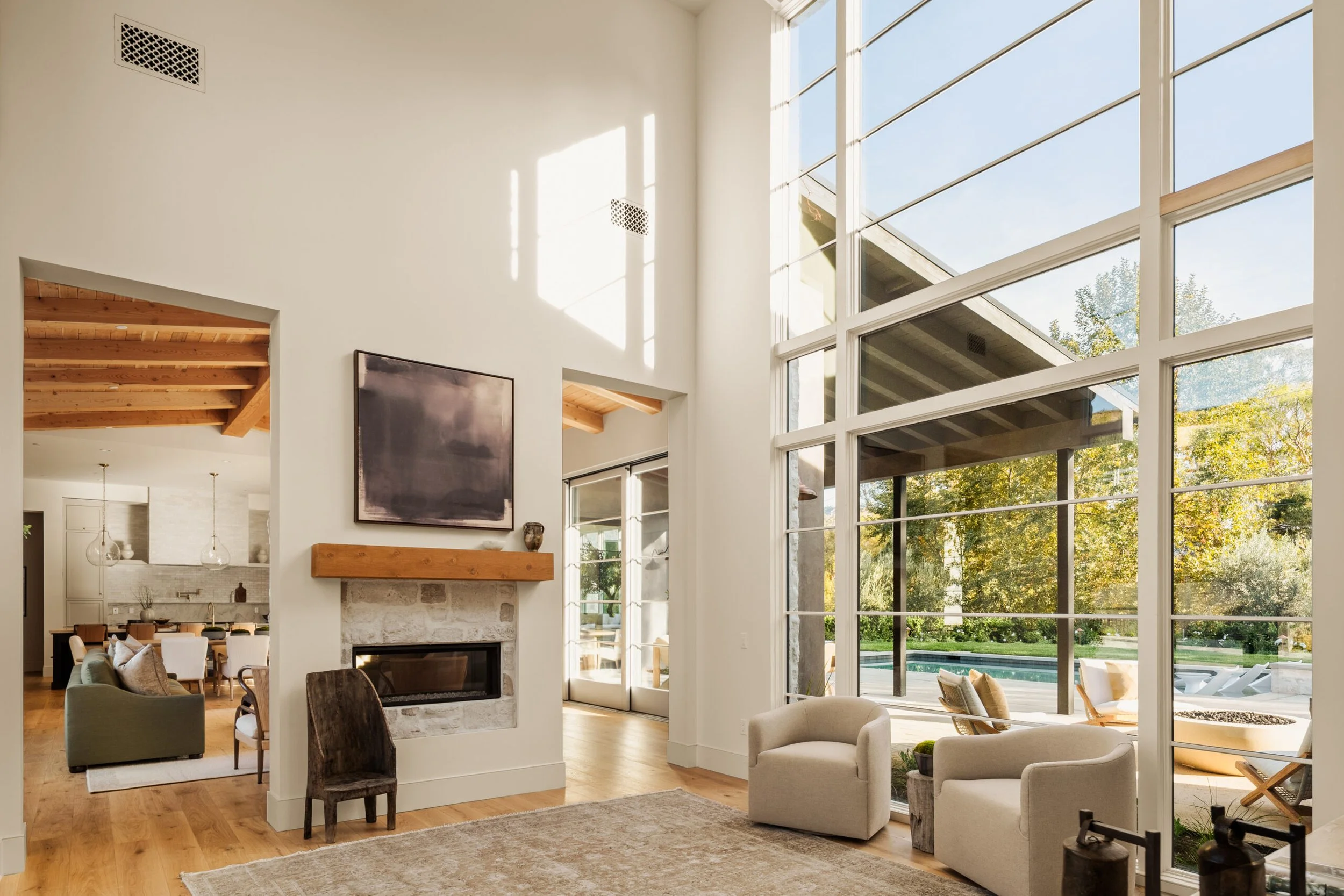 Bright living room with large floor-to-ceiling windows, modern furniture, a fireplace with a wooden mantel, and views of a backyard with a pool.