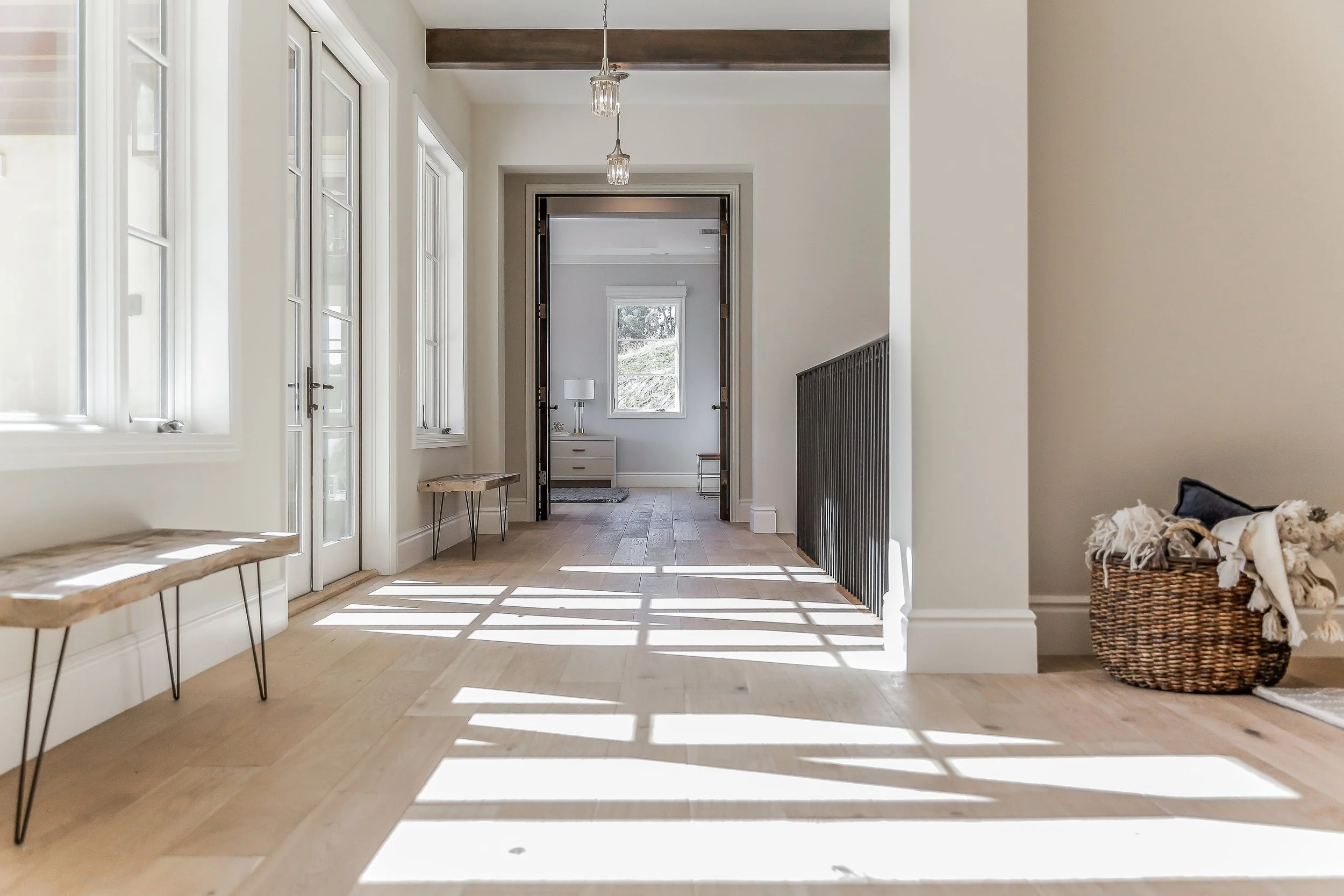 Bright entryway with multiple windows casting shadows on light hardwood floor, white walls, black handrail, and baskets with blankets on the right.