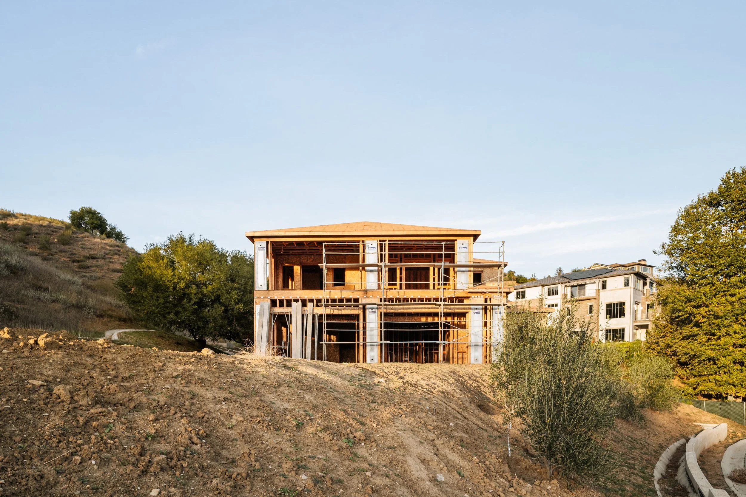 A two-story house under construction on a dirt hill, surrounded by trees and nearby residential buildings, with a clear sky overhead.