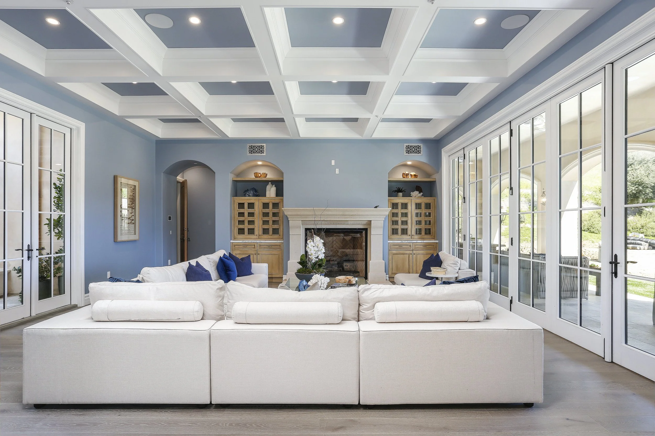Living room with light blue walls, white sectional sofa with blue pillows, large glass doors, built-in wooden cabinets, fireplace, and coffered ceiling.