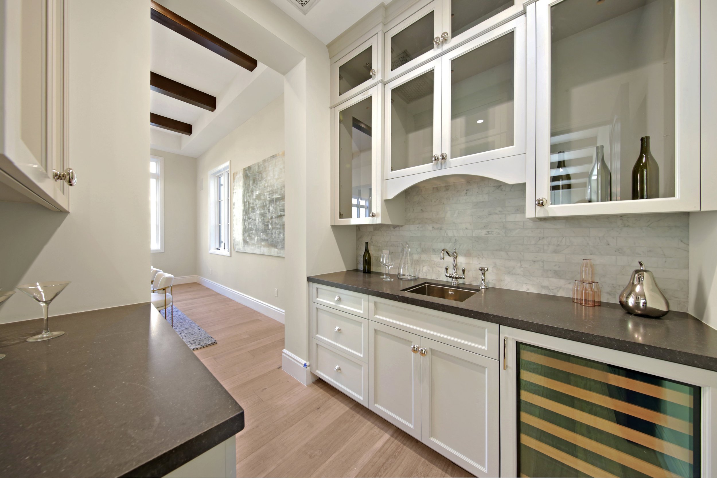 View of a modern kitchen with white cabinets, dark countertops, and a wine cooler, leading into a bright dining area with wooden floors and artwork on the wall.