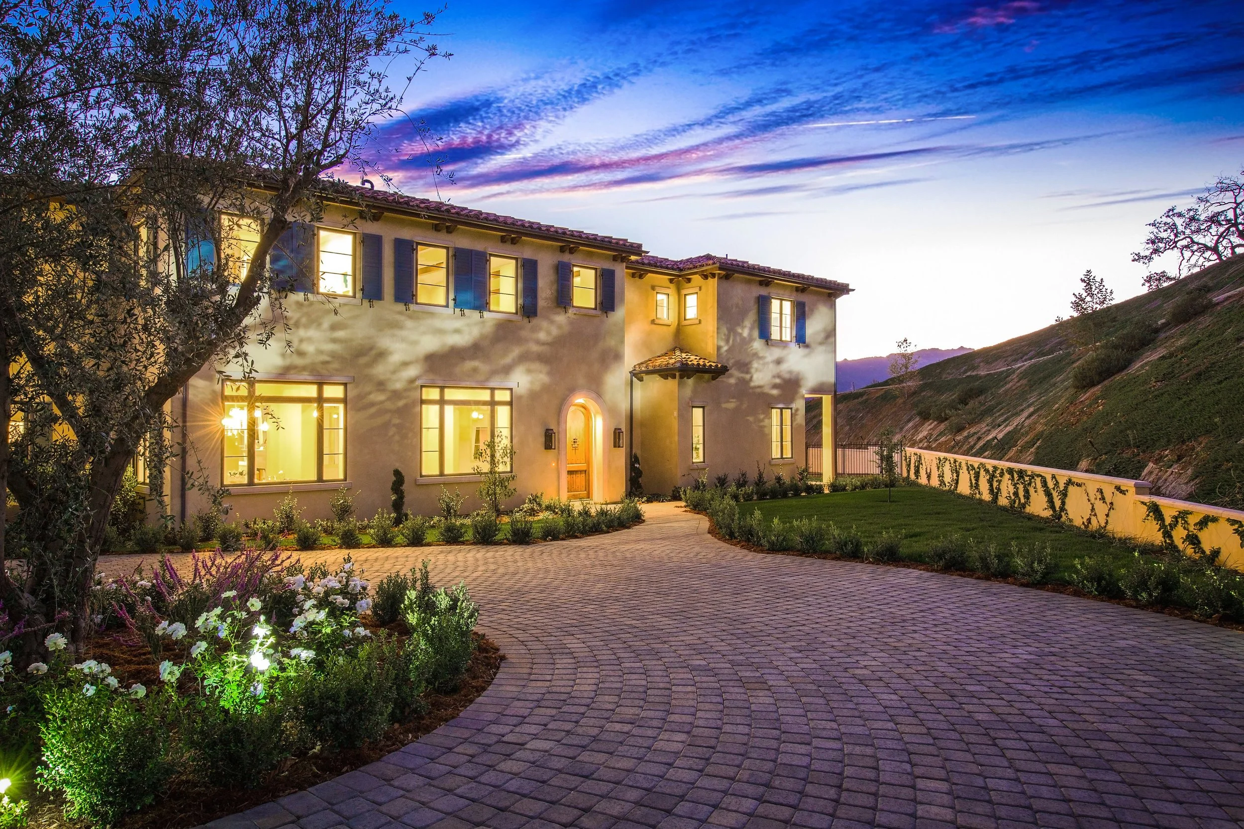 A large, two-story house illuminated at dusk with warm interior lighting, a curved paved driveway, and landscaped garden in front.
